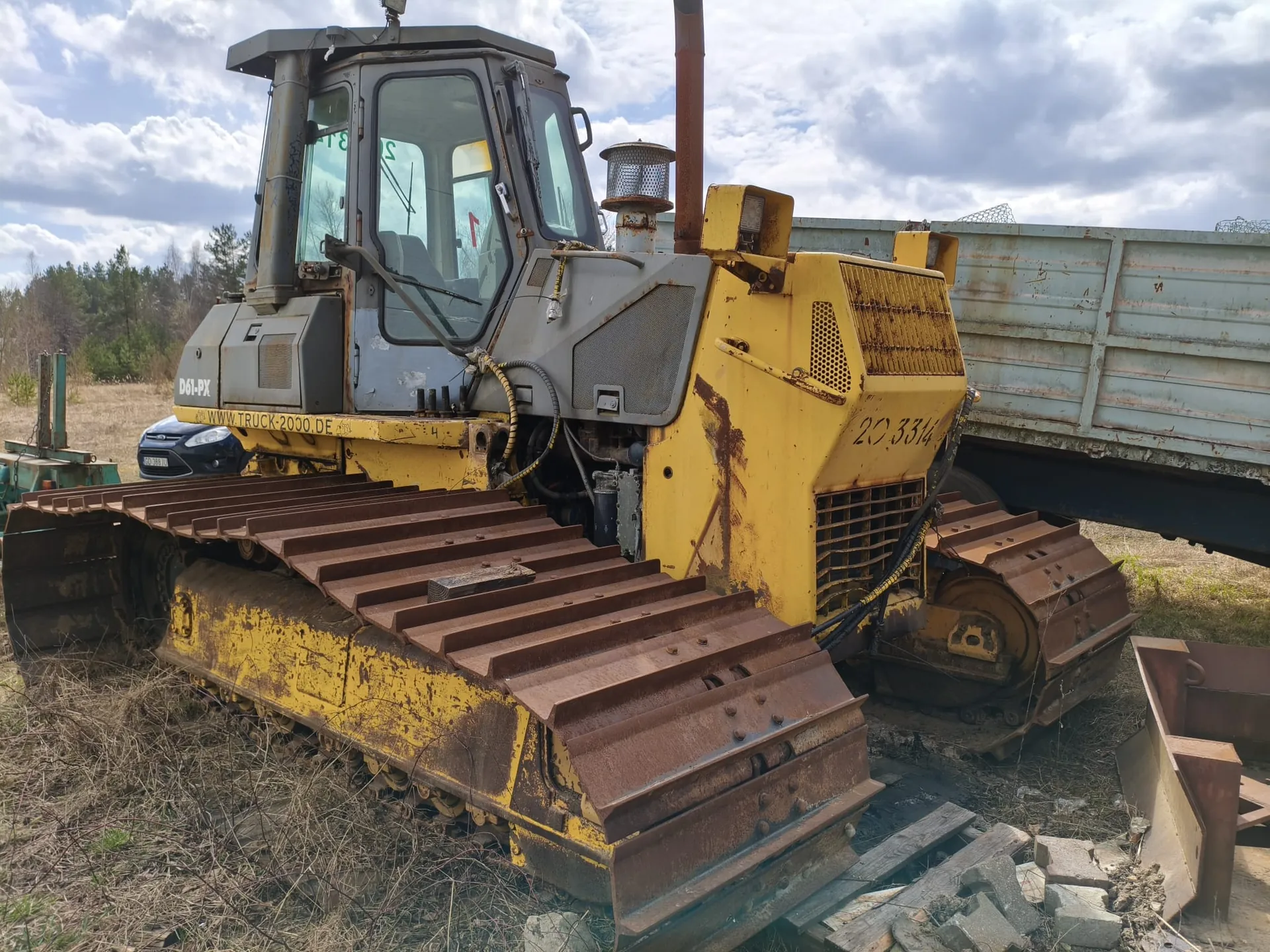 Stary, zardzewiały buldożer o kolorze żółto-szarym z gąsienicami, na którym widoczne są napisy: D61-PX, WWW.TRUCK-2000.DE oraz 20 3314. W tle znajduje się niebieski samochód, zielony zbiornik, drzewa i chmurne niebo. Na ziemi leżą kawałki cegieł i desek. Maszyna stoi na trawiastym terenie z suchą trawą.