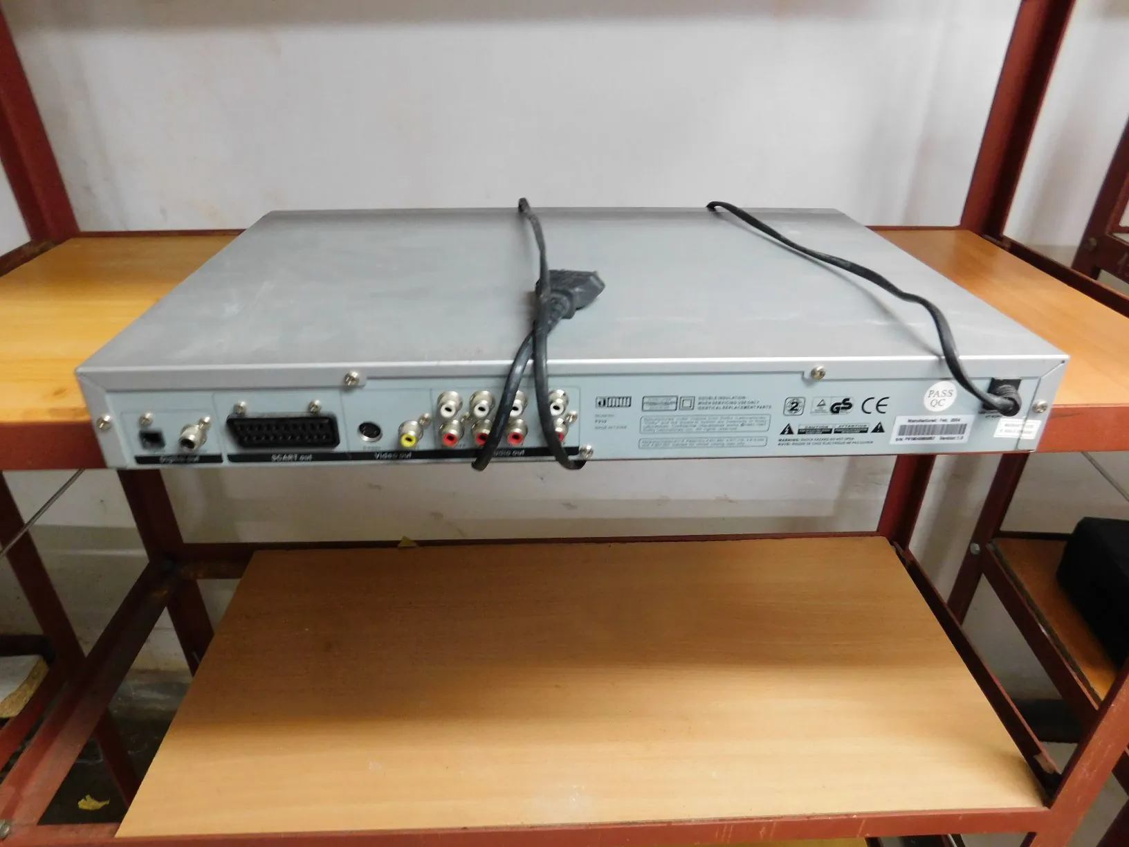Srebrne urządzenie elektroniczne leżące na drewnianej półce. Widoczny jest tylny panel z różnymi złączami, w tym gniazdo SCART, gniazda RCA (żółte, białe, czerwone) oraz gniazda typu BNC. Na obudowie znajduje się tabliczka znamionowa z certyfikatami CE i GS oraz naklejka 'PASS QC'. Do urządzenia podłączony jest czarny kabel zasilający.
