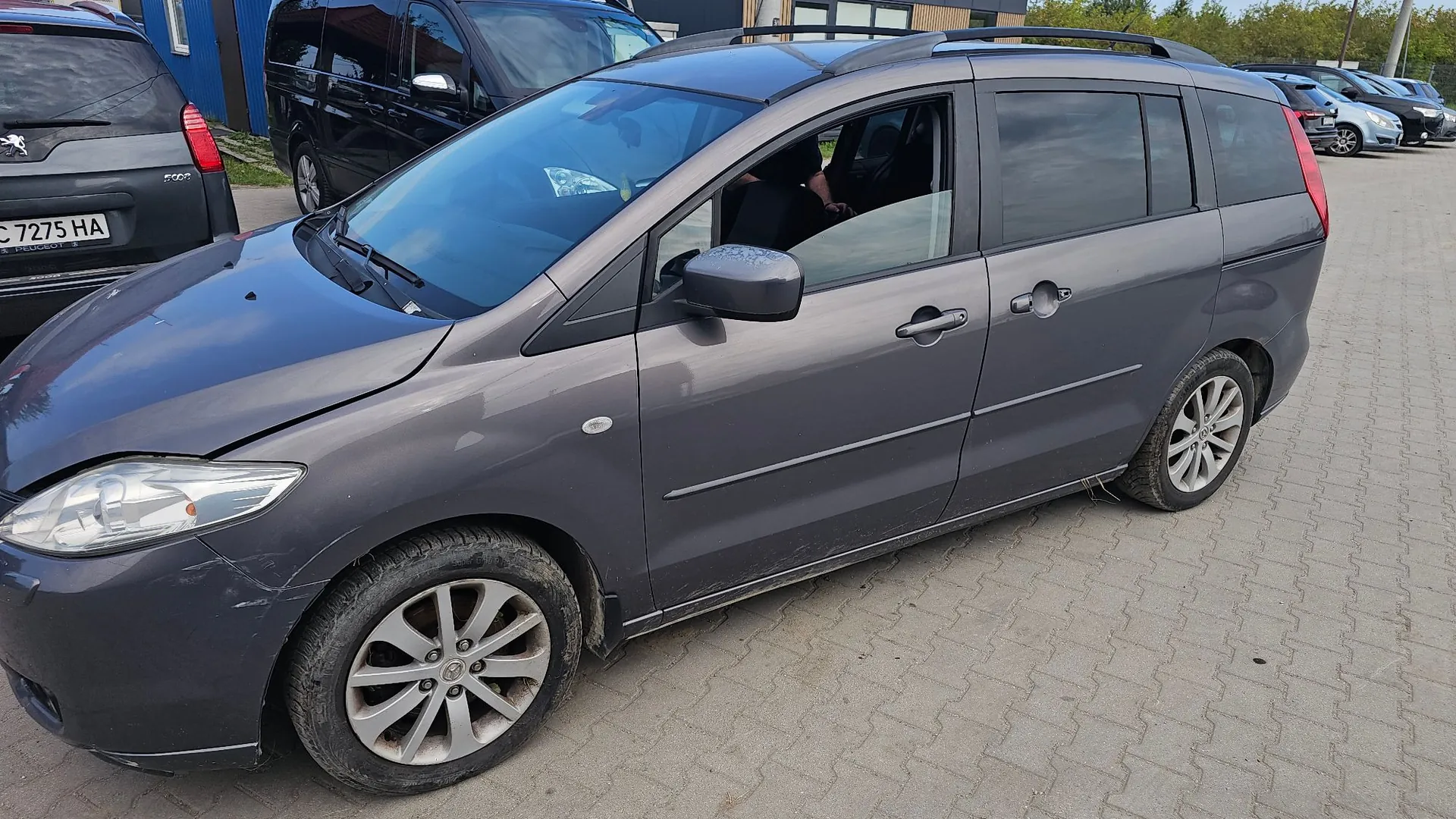 Szary samochód typu minivan zaparkowany na kostce brukowej. Pojazd jest widoczny z profilu od strony kierowcy. W tle widoczne są inne zaparkowane samochody oraz fragment niebieskiego budynku. Na samochodzie po lewej stronie widoczna jest tablica rejestracyjna o numerze C 7275 HA.