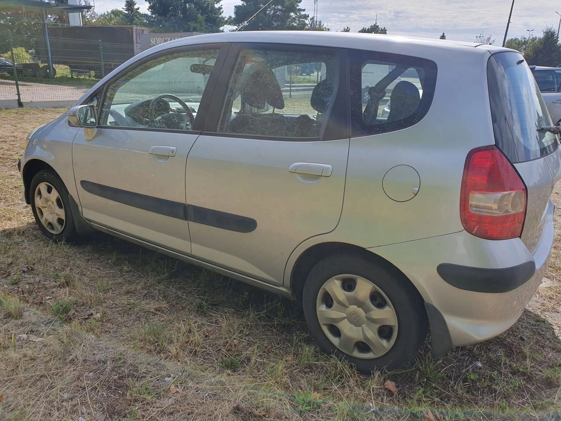 Srebrny samochód osobowy typu hatchback zaparkowany na trawiastym poboczu. Widok z boku, od strony pasażera. Pojazd posiada czarne listwy ochronne na drzwiach i zderzakach oraz srebrne kołpaki na kołach.