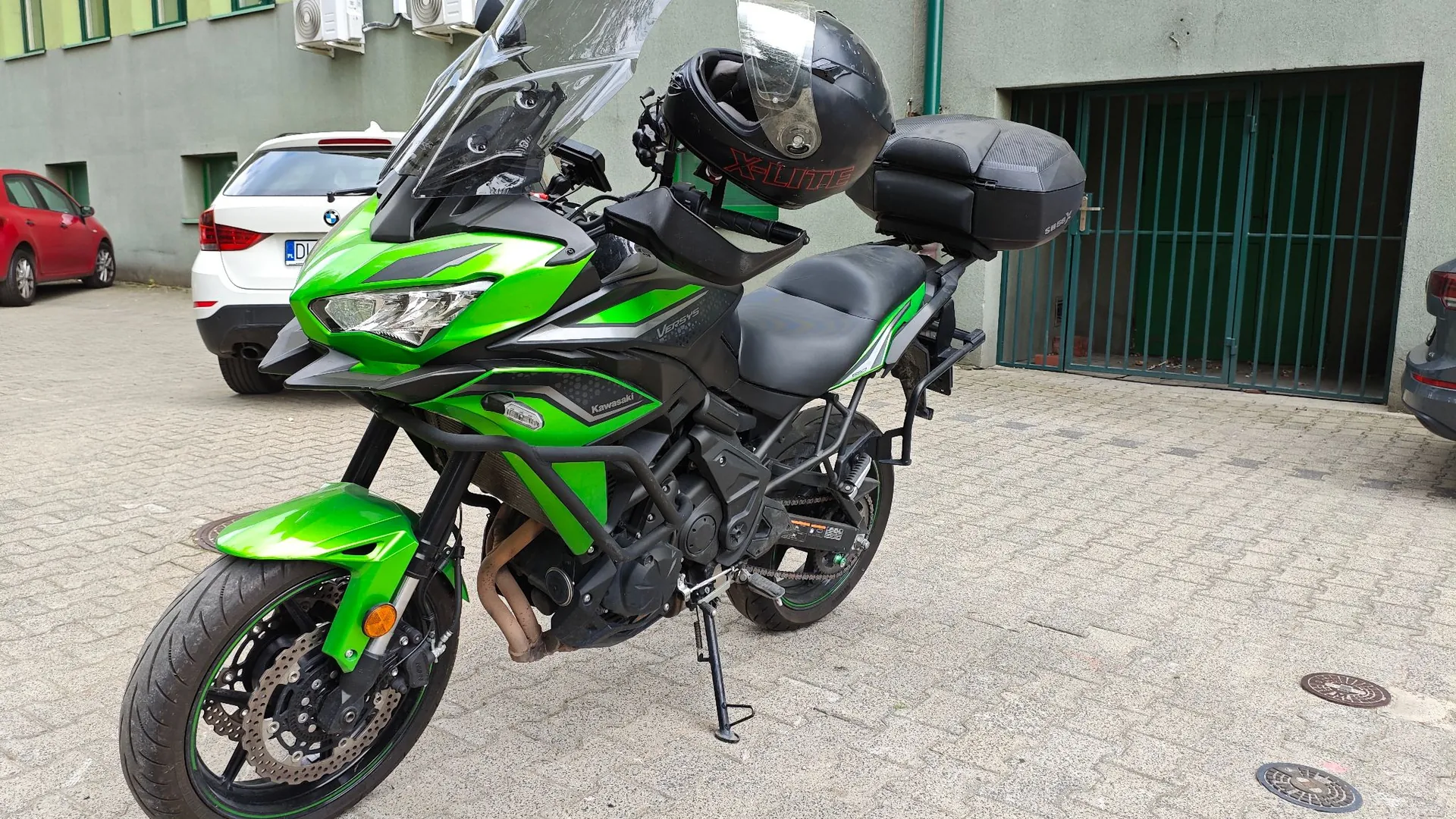 Zielony motocykl marki Kawasaki zaparkowany na kostce brukowej przed budynkiem. Motocykl jest wyposażony w wysoką szybę, czarny kufer tylny oraz czarny kask położony na siedzeniu. W tle widoczny jest biały samochód BMW oraz fragment czerwonego samochodu, a także zielona brama w ścianie budynku.