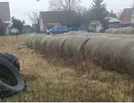 Pole z rzędem dużych, cylindrycznych balów siana ułożonych poziomo. Po lewej stronie widoczna duża opona. W tle domy, drzewa oraz niebieski samochód zaparkowany blisko budynków. Trawa na polu sucha, brązowa. Niebo zachmurzone.