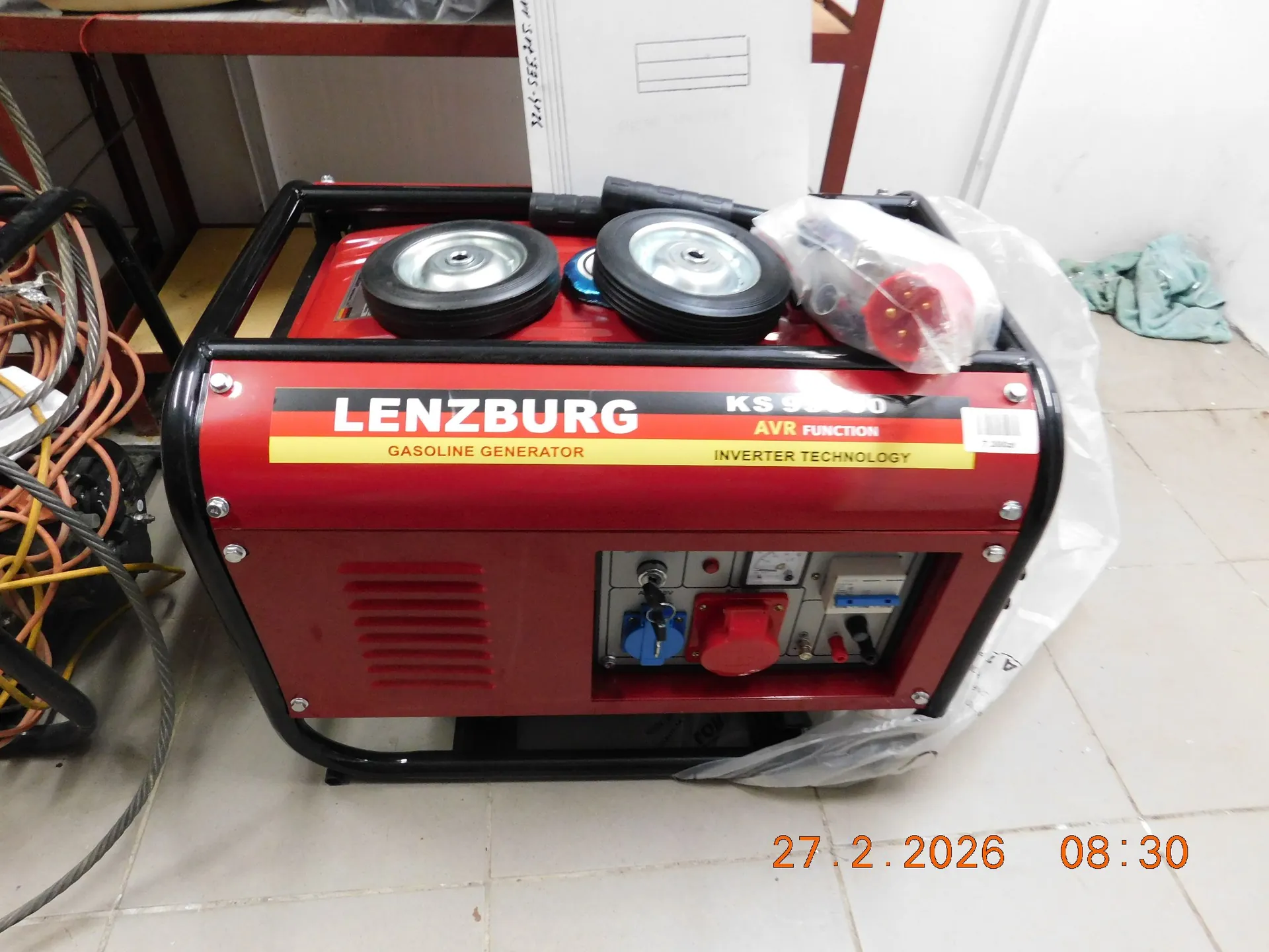 Czerwony agregat prądotwórczy marki LENZBURG, model KS 9500, z napisem 'GASOLINE GENERATOR', 'AVR FUNCTION' oraz 'INVERTER TECHNOLOGY'. Urządzenie posiada czarną metalową ramę ochronną, dwa czarne koła transportowe na górze oraz panel sterowania z gniazdami elektrycznymi i przełącznikami z przodu. Agregat stoi na jasnej płytkowej podłodze, obok znajduje się plastikowa folia ochronna.