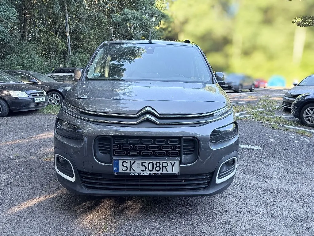 Szary samochód marki Citroën zaparkowany przodem do obiektywu na asfaltowym parkingu. Pojazd posiada polską tablicę rejestracyjną o numerze SK 508RY. W tle widoczne są inne zaparkowane samochody oraz gęste, zielone drzewa.