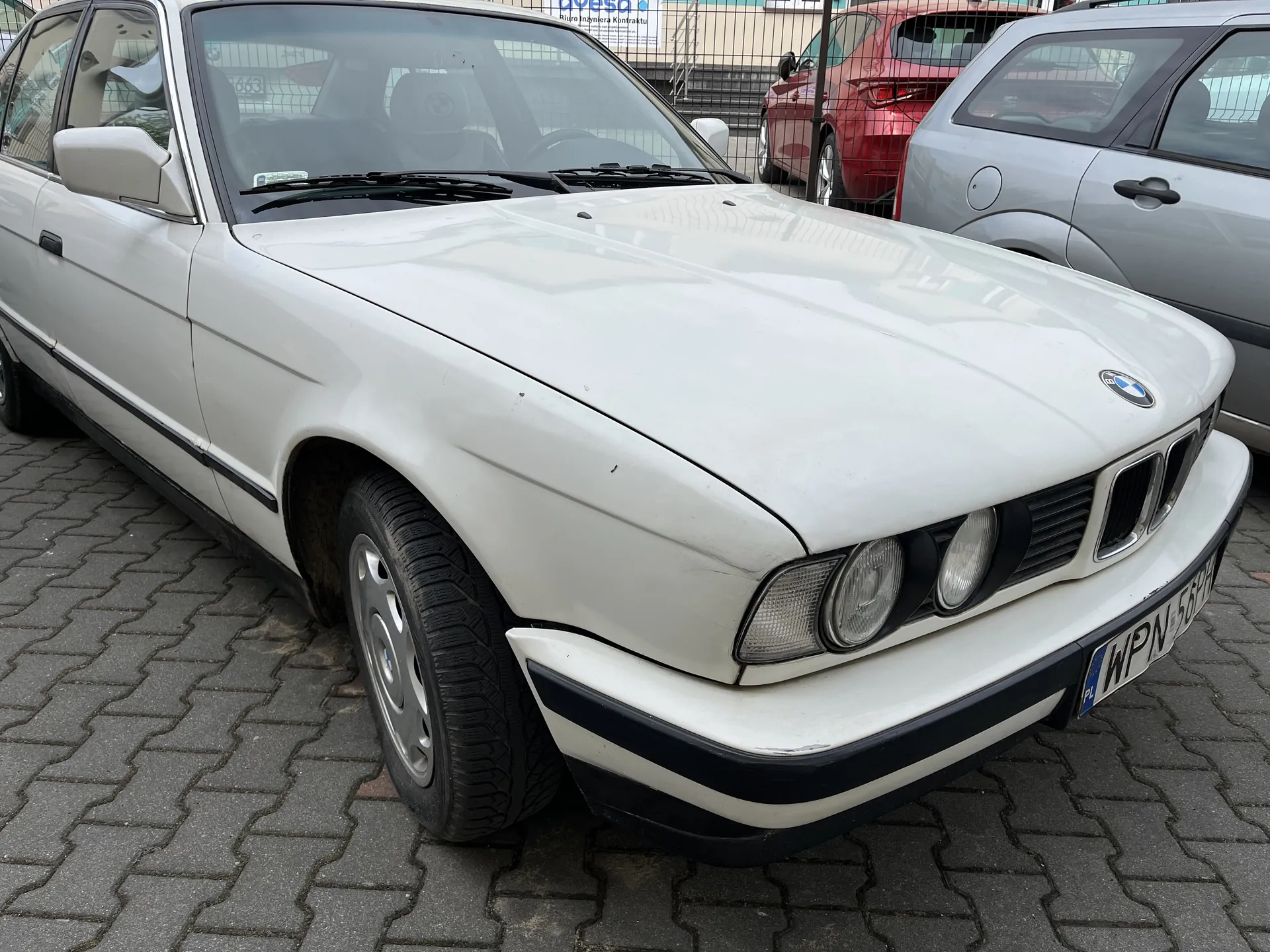Biały samochód marki BMW z tablicą rejestracyjną WPN 55H, zaparkowany na brukowanej powierzchni. Widoczne są przednie reflektory, charakterystyczna atrapa chłodnicy z logo BMW oraz czarna listwa biegnąca wzdłuż pasa bocznego. W tle stoją czerwony i srebrny samochód, a za nimi metalowe ogrodzenie i fragment budynku z niebieskim napisem. Tekst na tablicy rejestracyjnej jest czytelny.