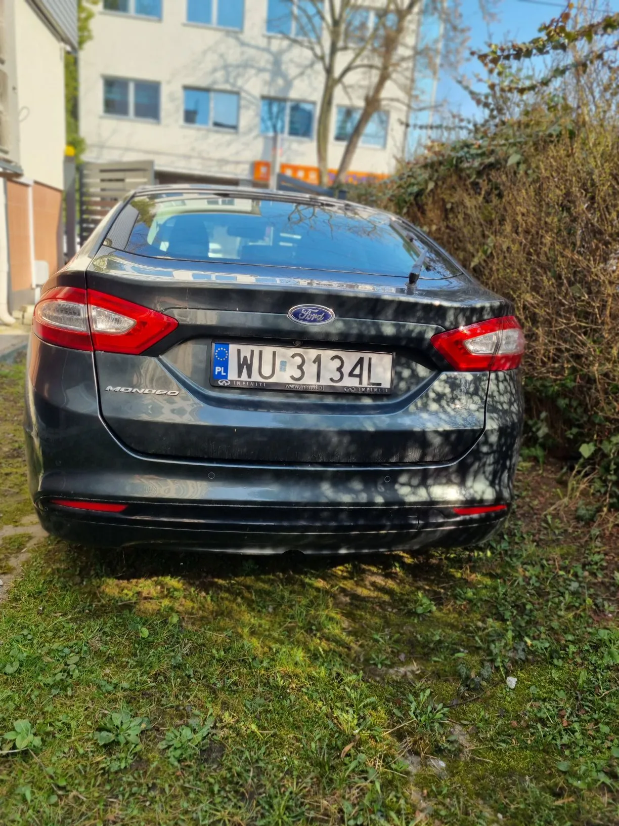 Czarny samochód Ford Mondeo widziany od tyłu z tablicą rejestracyjną WU 3134L (PL). W tle budynek z oknami, drzewa bez liści i trawa. Samochód zaparkowany na trawie.