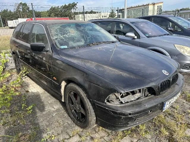 Czarny samochód marki BMW w nadwoziu kombi, zaparkowany na betonowym placu. Pojazd jest w złym stanie technicznym: przednia szyba jest mocno rozbita, brakuje reflektora w prawym przednim kloszu, a lakier na masce jest zmatowiony i zabrudzony. W tle widoczne są inne zaparkowane samochody, ogrodzenie z siatki oraz budynek przemysłowy.
