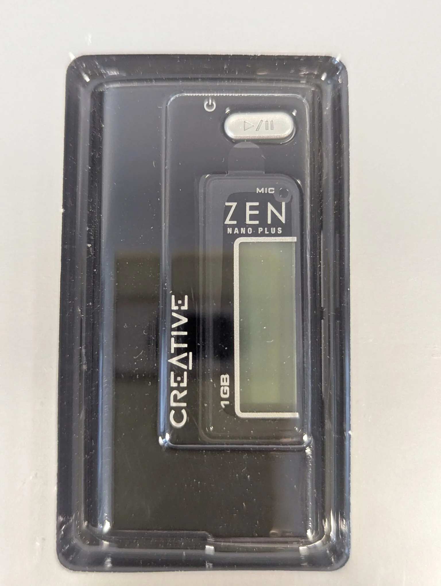 Czarny odtwarzacz MP3 marki Creative Zen Nano Plus o pojemności 1GB, zapakowany w przezroczyste plastikowe opakowanie. Na urządzeniu widoczny jest napis 'CREATIVE' pionowo po lewej stronie, napis 'ZEN NANO PLUS' nad małym ekranem LCD oraz napis '1GB' pod ekranem. Na górze znajduje się przycisk odtwarzania/pauzy oraz symbol zasilania.