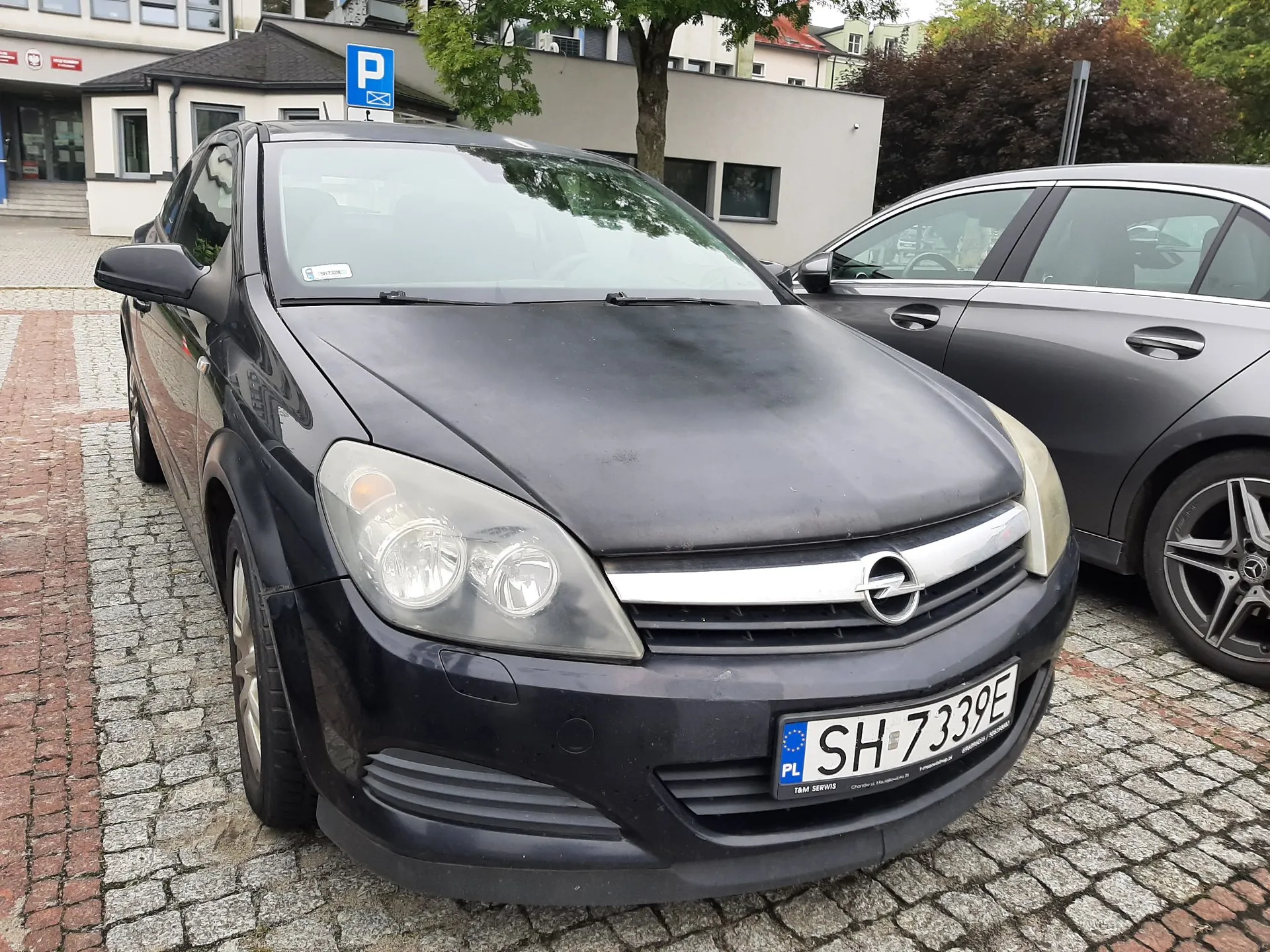 Czarny samochód marki Opel zaparkowany na kostce brukowej przed jasnym budynkiem. Widoczna jest przednia część pojazdu z tablicą rejestracyjną o numerze SH 7339E. Obok stoi inny, szary samochód. W tle widać budynek z niebieskim znakiem parkowania 'P'.