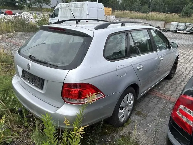 Srebrny samochód osobowy marki Volkswagen w nadwoziu kombi, zaparkowany tyłem do obiektywu na kostce brukowej. Pojazd posiada relingi dachowe i hak holowniczy. W tle widoczny jest biały furgon oraz teren przemysłowy z hałdami materiałów.