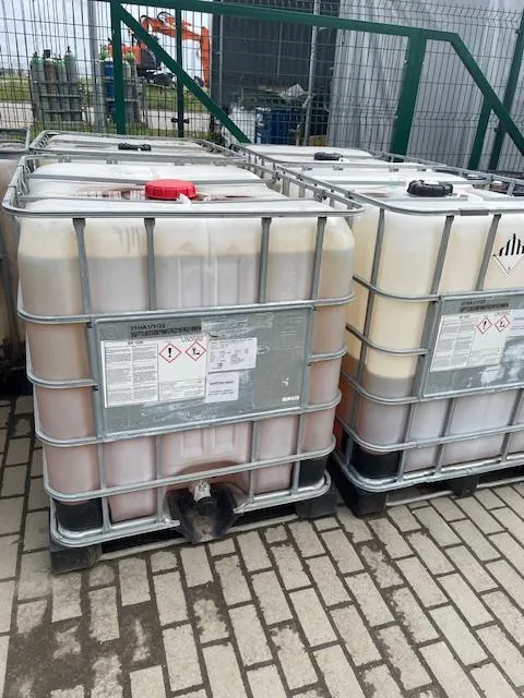 Kilka białych zbiorników IBC w metalowych klatkach, ustawionych obok siebie na kostce brukowej. Na pierwszym zbiorniku widoczna jest czerwona zakrętka oraz etykieta ostrzegawcza z piktogramami zagrożeń i tekstem. W tle znajduje się zielony płot z siatki oraz fragment pomarańczowej maszyny budowlanej.