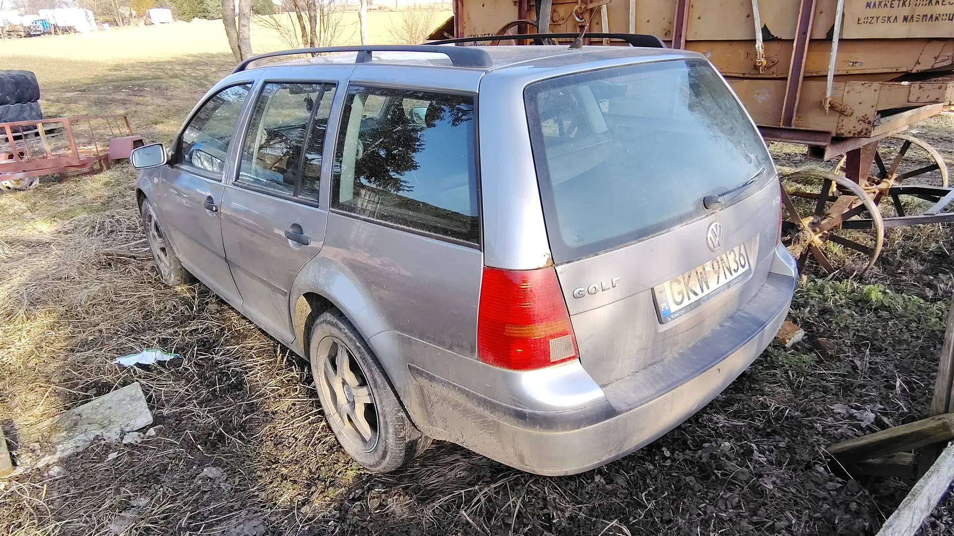 Srebrny samochód osobowy typu kombi Volkswagen Golf z tablicą rejestracyjną PL GKW 9N36, zaparkowany na trawiastym i błotnistym terenie. W tle widoczny stary, rdzewiejący pojazd rolniczy z częściowo czytelnym napisem 'NAKRETKI DOKRE...' oraz 'LOZYSKA NASHARO...', stos opon, czerwona metalowa konstrukcja i drzewa. Samochód posiada dachówkę i jest zabrudzony. Na ziemi leżą suche trawy i śmieci.
