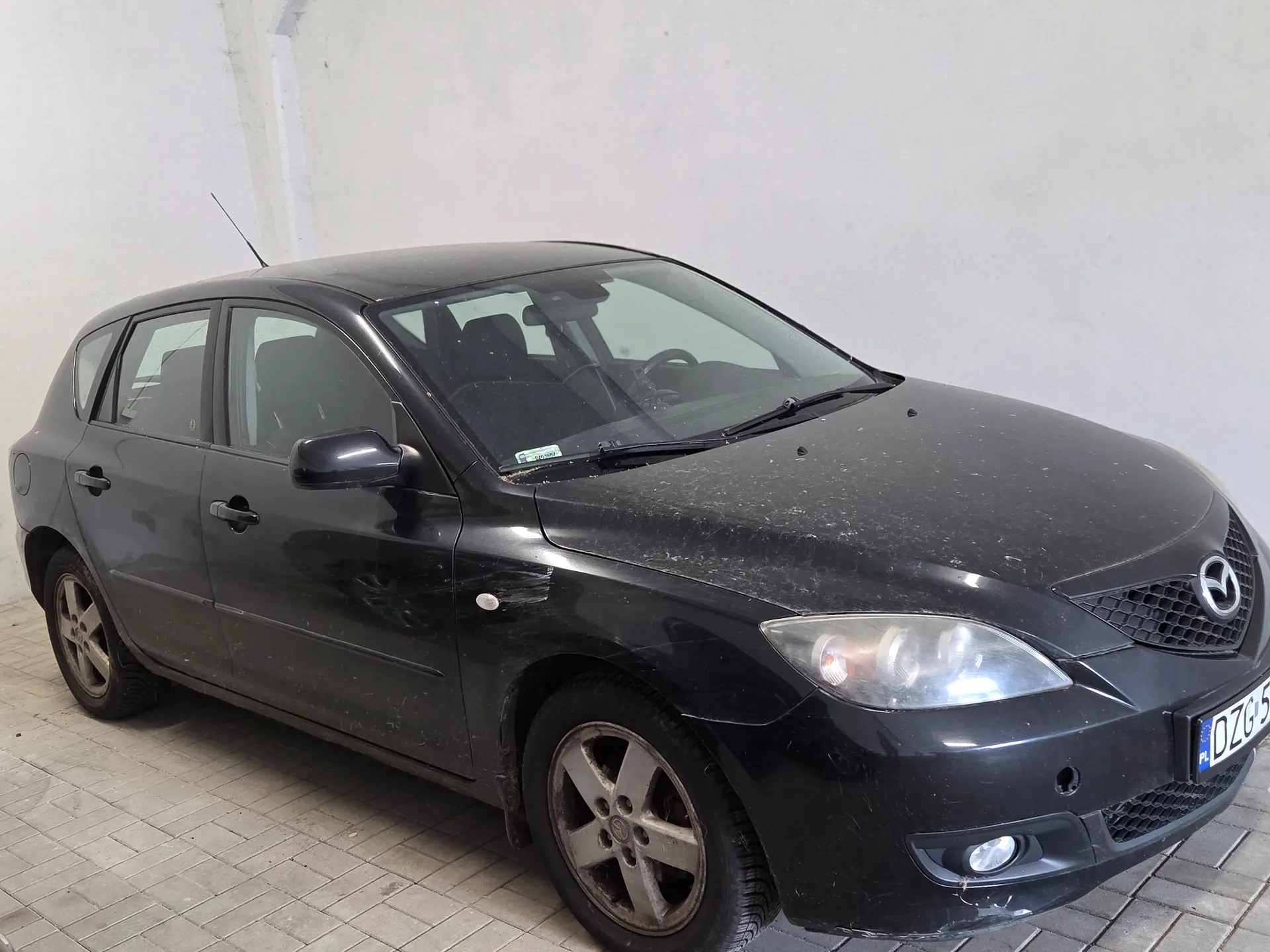 Czarny samochód marki Mazda w nadwoziu hatchback, zaparkowany na kostce brukowej przed białą ścianą. Pojazd jest zakurzony, widoczna jest przednia część auta z reflektorami, logo Mazdy na masce oraz fragment tablicy rejestracyjnej z literami DZG.