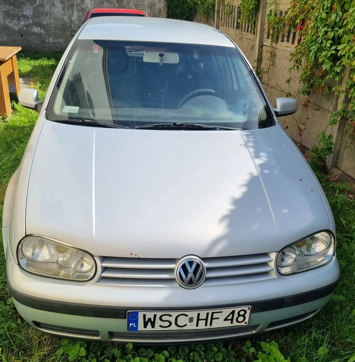 Srebrny samochód marki Volkswagen widziany od przodu, zaparkowany na trawie obok betonowego ogrodzenia porośniętego roślinnością. Na przednim zderzaku znajduje się polska tablica rejestracyjna o numerze WSC HF48.