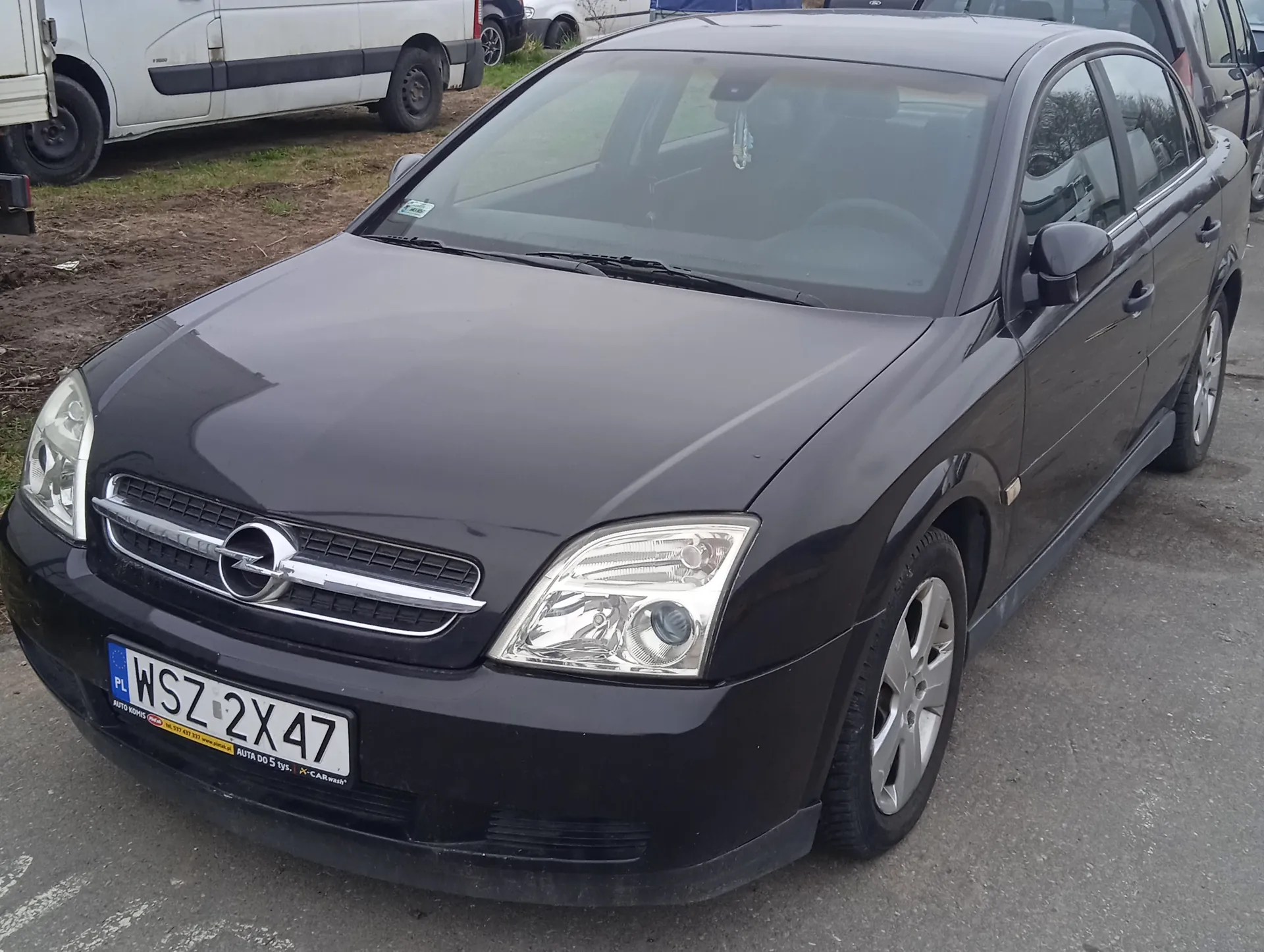 Czarny samochód marki Opel z tablicą rejestracyjną PL WSZ 2X47. Na tablicy widoczny żółty naklejka z napisem 'AUTO DO 5 tys. X-CAR.wash'. W tle biały furgon i trawa na brudnej ziemi. Samochód stoi na asfaltowej powierzchni, przednie światła i atrapa chłodnicy są widoczne.