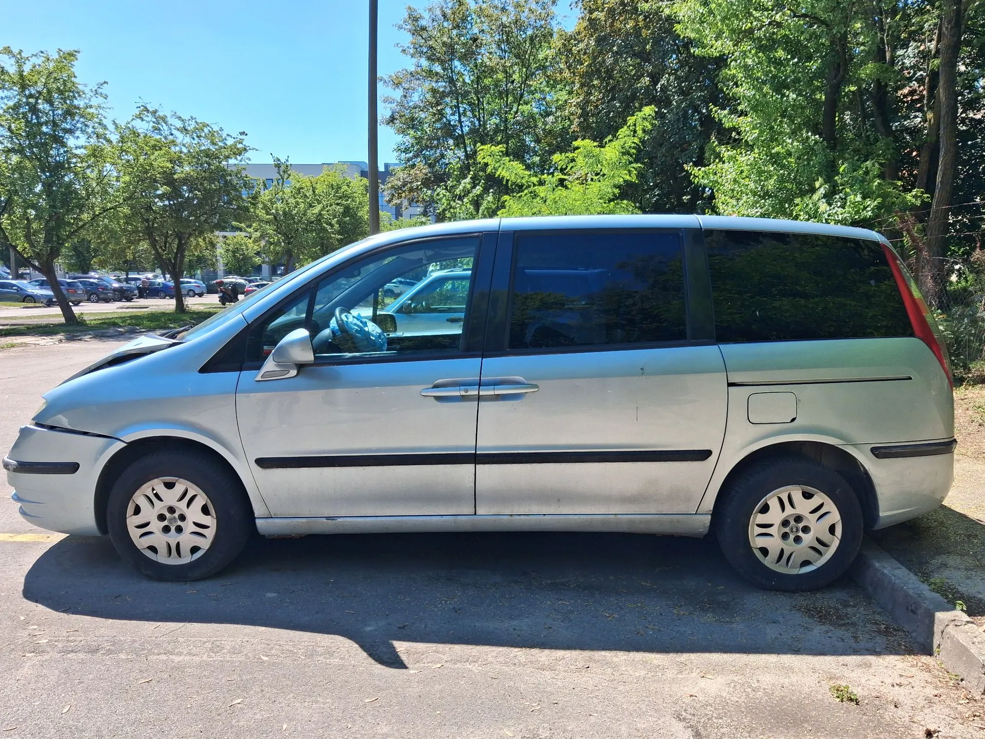 Srebrny samochód typu minivan zaparkowany bokiem na asfaltowym parkingu. W tle widoczne są zielone drzewa, inne zaparkowane pojazdy oraz fragment budynku pod jasnym, bezchmurnym niebem.