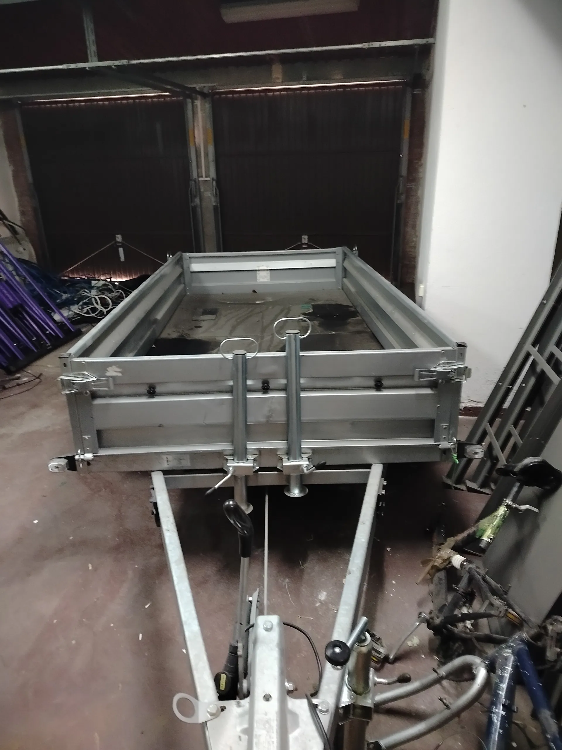 Srebrny metalowy przyczepa o prostokątnym kształcie z dwoma pionowymi słupkami z pierścieniami na górze. W tle ciemne bramy garażowe, po lewej stronie fioletowe konstrukcje i przewody, po prawej drabina oraz części rowerów. Podłoga z betonu z widocznymi zabrudzeniami. Całość w pomieszczeniu przypominającym garaż lub magazyn.