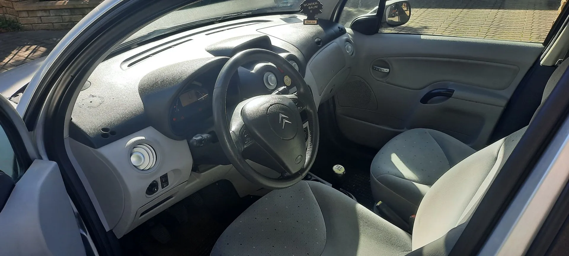 Wnętrze samochodu marki Citroën widziane z perspektywy otwartych drzwi kierowcy. Widoczna jest szara deska rozdzielcza, kierownica z logo Citroëna, dwa szare fotele materiałowe oraz panel sterowania po lewej stronie. Na górnej części deski rozdzielczej znajduje się mała ozdoba. Przez przednią szybę widoczny jest fragment podjazdu z kostki brukowej.