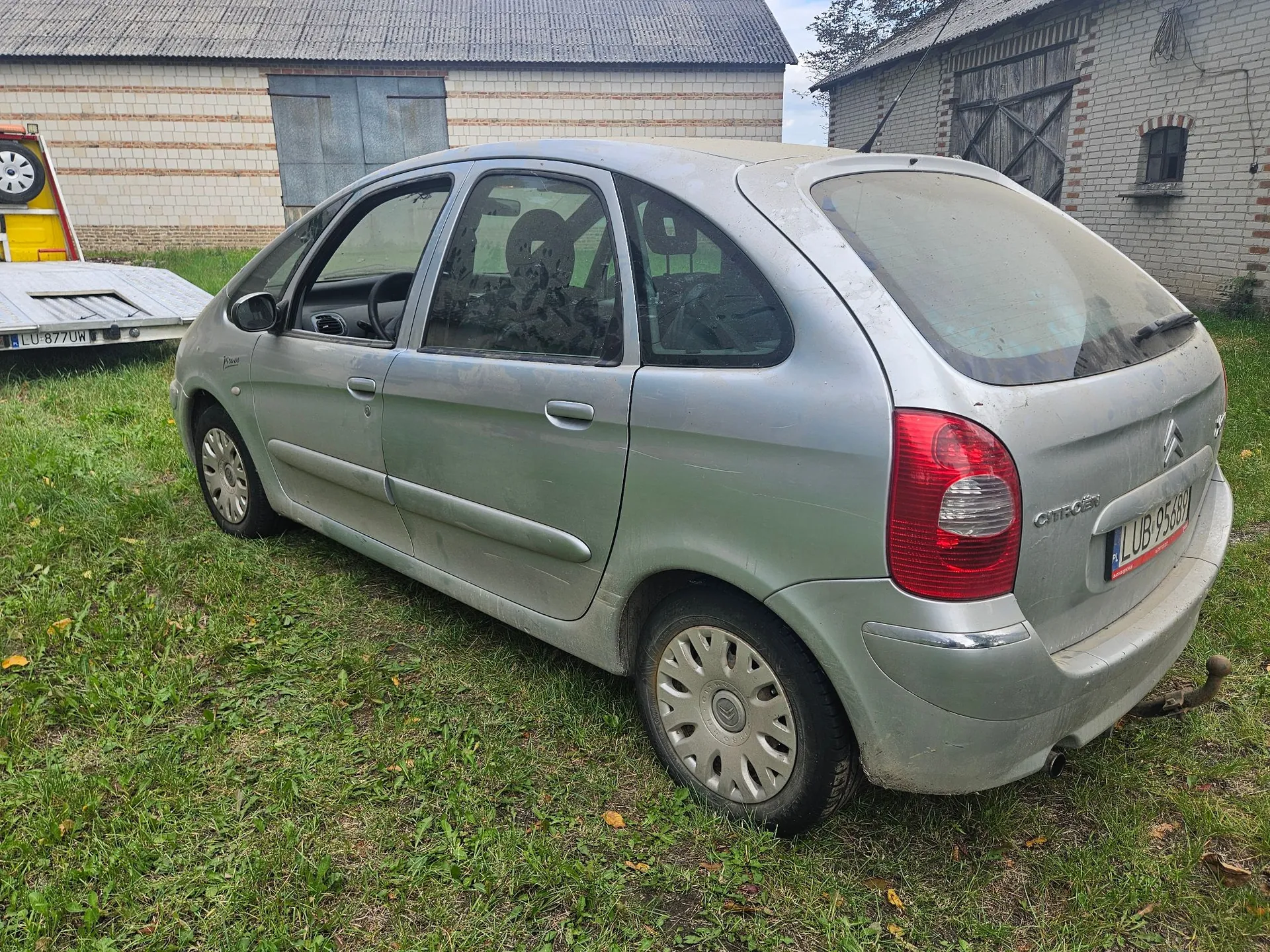 Srebrny samochód osobowy marki Citroën w nadwoziu hatchback, zaparkowany na trawie przed budynkami gospodarczymi. Pojazd jest widoczny z tyłu i z boku, posiada tablicę rejestracyjną o numerze LUB 956GY. W tle widoczna jest biała stodoła z szarym dachem oraz fragment żółtej przyczepy z tablicą LU 8770W.