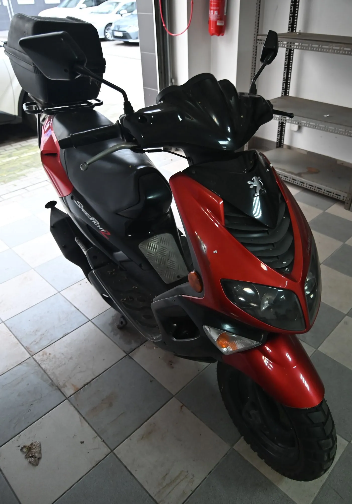 Czerwony skuter marki Peugeot z czarnymi elementami, zaparkowany w garażu na jasnej, kafelkowej podłodze. Pojazd posiada czarny kufer zamykany z tyłu, czarną kanapę oraz czarną owiewkę kierownicy. Na boku skutera widnieje napis 'Speedfight 2'. W tle widoczne są metalowe regały magazynowe oraz fragmenty białych samochodów za otwartą bramą garażową.