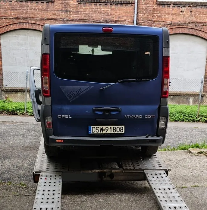Niebieski samochód dostawczy marki Opel Vivaro CDT, zaparkowany tyłem na platformie lawety. Pojazd posiada polską tablicę rejestracyjną o numerze DSW 91808. Na tylnej szybie znajduje się niebieska naklejka z napisem „Gebra MILDE”. W tle widoczny jest ceglany budynek z dwiema białymi bramami garażowymi.