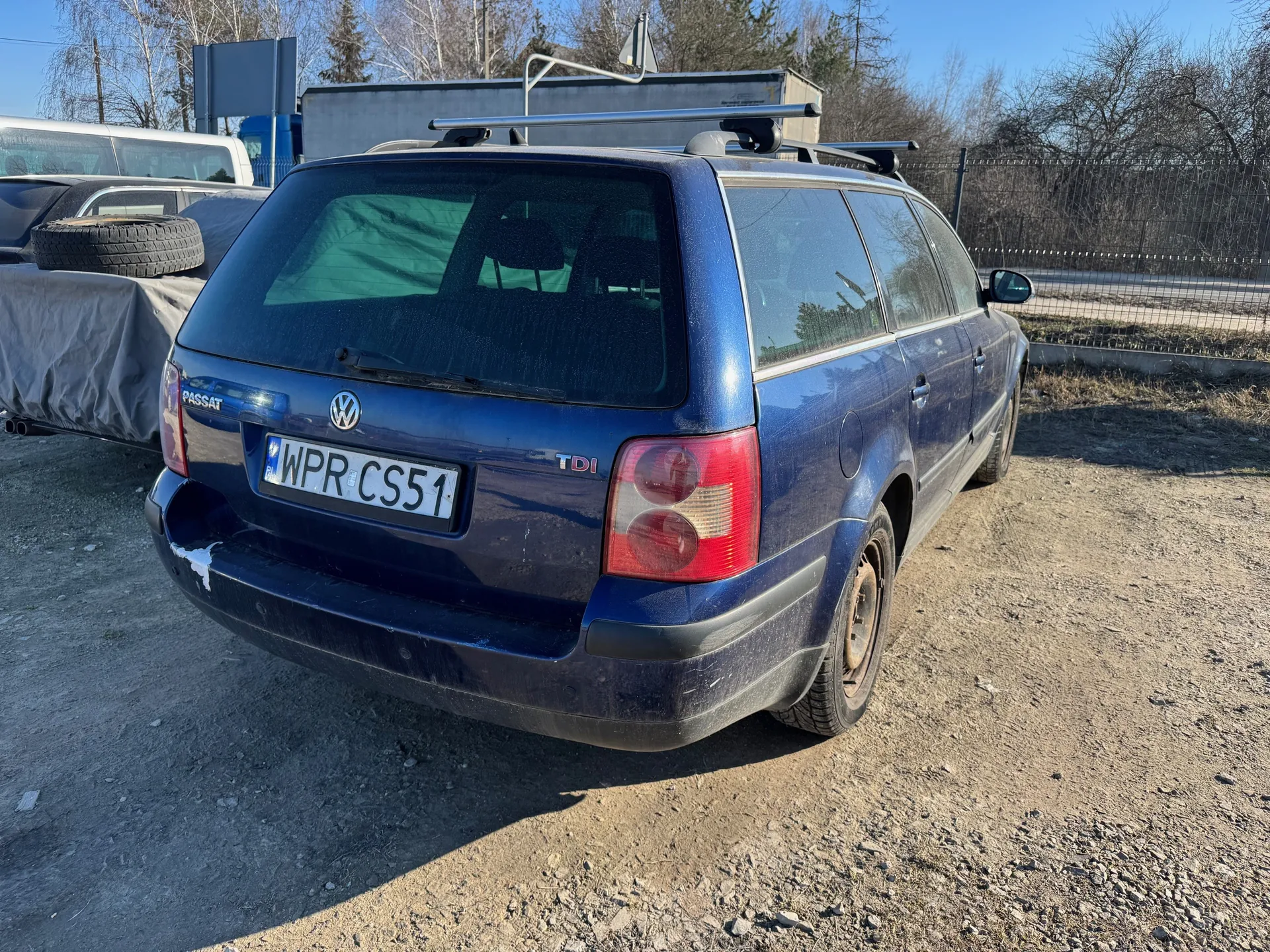 Niebieski samochód osobowy Volkswagen Passat kombi z widocznym logo VW na tylnej szybie, napisem PASSAT po lewej stronie bagażnika i oznaczeniem TDI po prawej. Tablica rejestracyjna WPR CS51 umieszczona na środku bagażnika. Samochód wyposażony w dachowy bagażnik. W tle: częściowo zakryty szarą pokrywą pojazd z oponą na dachu, metalowe ogrodzenie, suche drzewa, niebo oraz nierówna, żwirowa powierzchnia podłogi. Na tylnym szybie widoczne odbicia drzew i budynku.