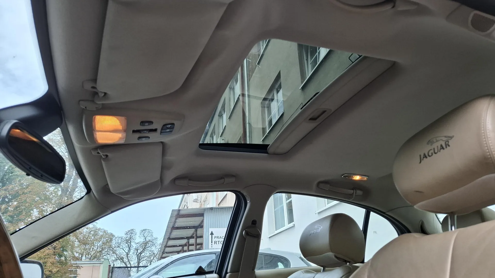 Widok z wnętrza samochodu marki Jaguar, skierowany w stronę dachu. Widoczny jest otwarty dach panoramiczny, przez który widać fasadę budynku. Wnętrze auta jest w kolorze beżowym, z widocznym zagłówkiem z napisem 'JAGUAR'. Na podsufitce znajdują się lampki oświetlenia wnętrza oraz panel sterowania.