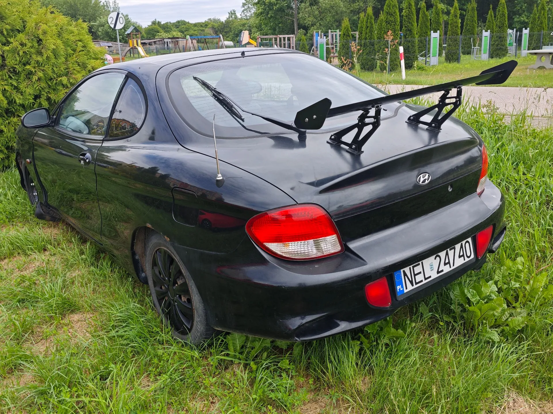 Czarny samochód marki Hyundai w nadwoziu coupe, zaparkowany na trawie. Pojazd posiada duży, czarny spojler zamontowany na tylnej klapie. Widoczna jest tablica rejestracyjna o treści NEL 24740 z oznaczeniem PL. W tle widać zieleń, krzewy oraz fragment placu zabaw.