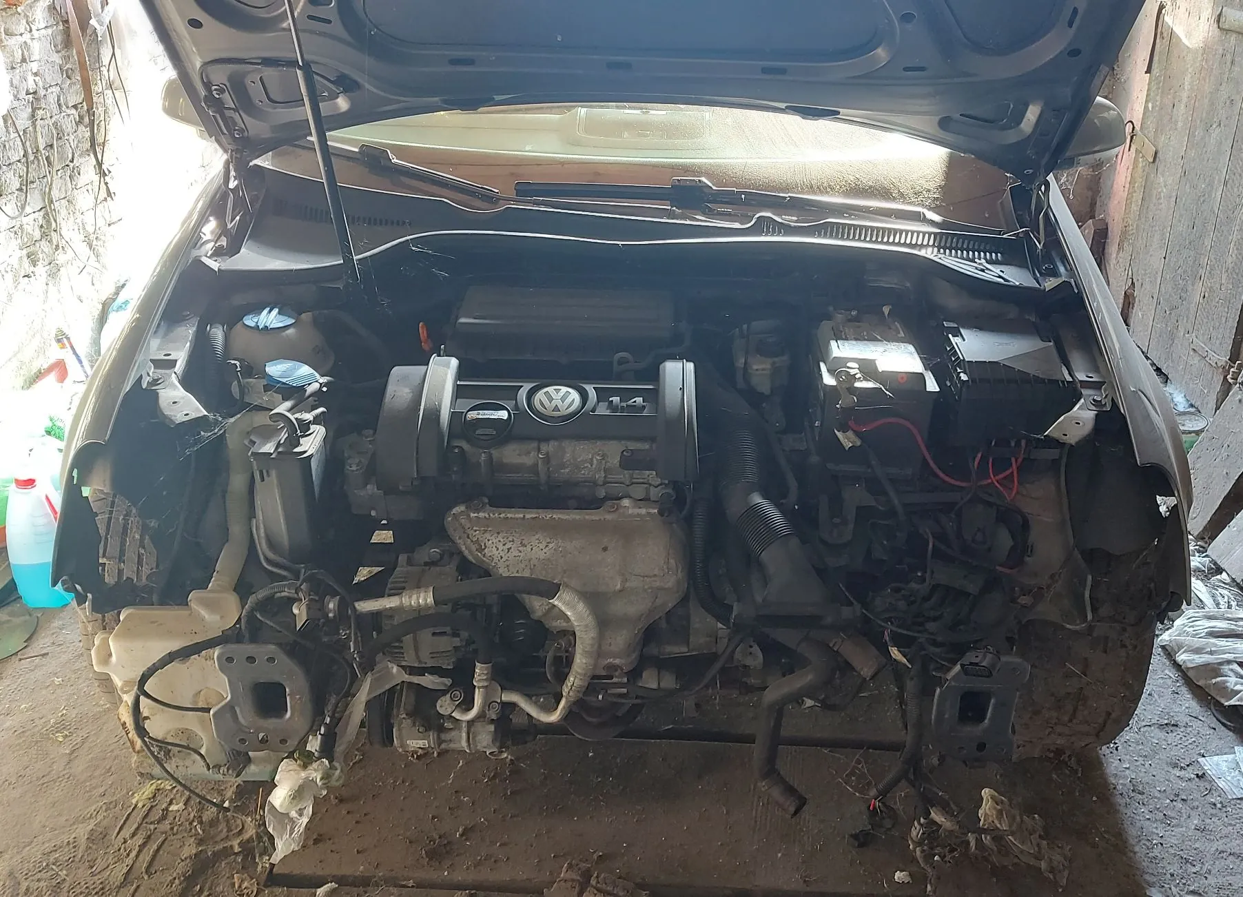 Widok na komorę silnika samochodu marki Volkswagen z otwartą maską. Samochód jest w stanie rozbiórki lub po kolizji, brakuje przedniego zderzaka oraz części elementów nadwozia. Widoczny jest silnik z logo VW na osłonie, akumulator po prawej stronie oraz różne przewody i elementy mechaniczne.