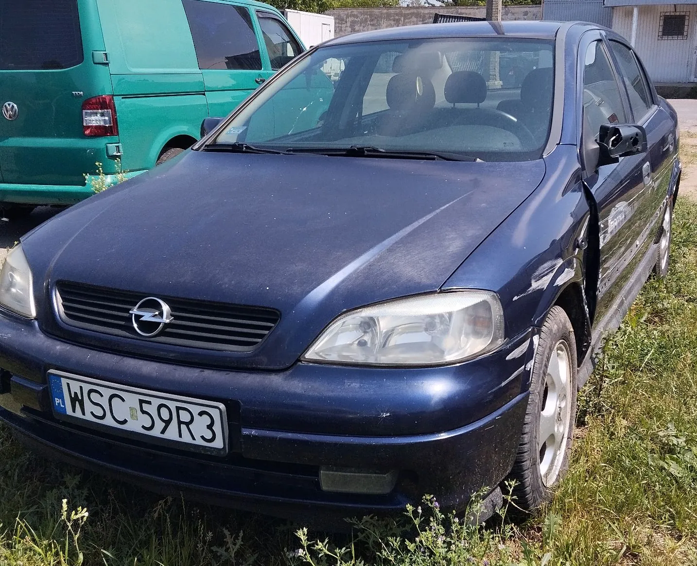 Granatowy samochód osobowy marki Opel zaparkowany na trawie. Pojazd ma widoczne uszkodzenia karoserii po prawej stronie (stłuczone drzwi pasażera). Na przednim zderzaku znajduje się polska tablica rejestracyjna o numerze WSC 59R3. W tle widoczny jest fragment zielonego vana marki Volkswagen.