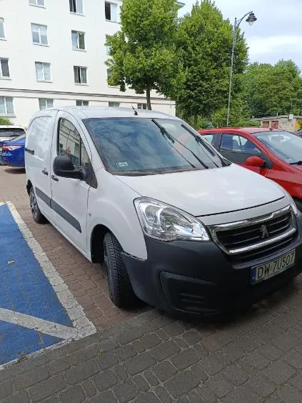 Biały samochód dostawczy marki Peugeot zaparkowany na kostce brukowej. Pojazd ma czarny zderzak przedni i tablicę rejestracyjną o numerze DW 70507. Obok stoi czerwony samochód osobowy, a w tle widoczny jest biały budynek mieszkalny z oknami oraz zielone drzewa.