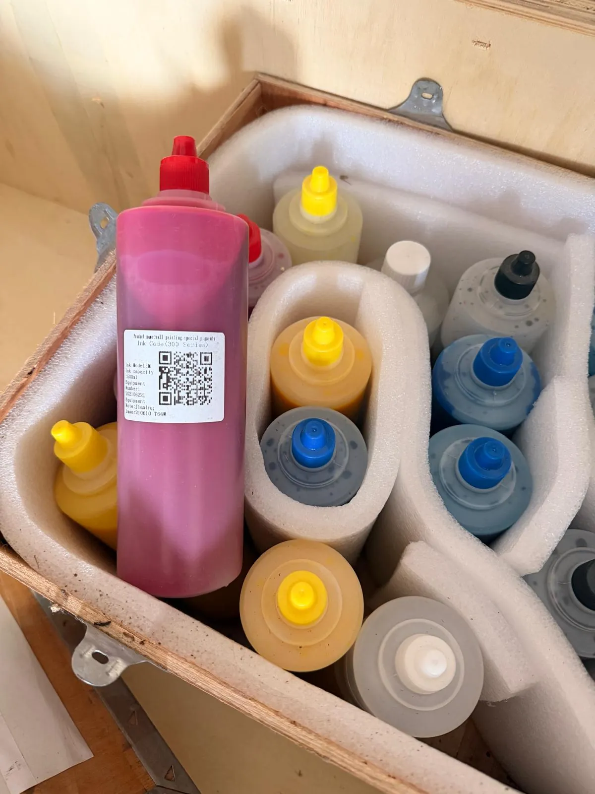 Kilka plastikowych butelek z farbami lub tuszami w różnych kolorach (różowy, żółty, niebieski, szary), umieszczonych w białym opakowaniu z pianki styropianowej wewnątrz drewnianej skrzyni. Najbardziej widoczna jest duża, różowa butelka z etykietą zawierającą kod QR i tekst: 'Ink Code: 1302 (pink)'.