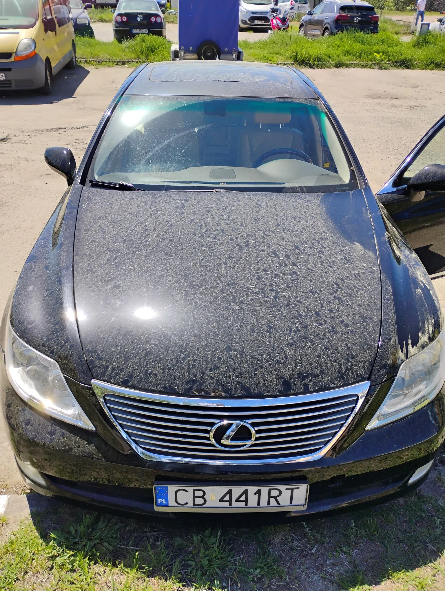 Czarny samochód marki Lexus zaparkowany na nieutwardzonym terenie. Widoczny jest przód pojazdu z maską pokrytą kurzem, charakterystycznym grillem z logo marki oraz tablicą rejestracyjną o numerze CB 441RT. W tle widoczne są inne zaparkowane samochody oraz trawa.