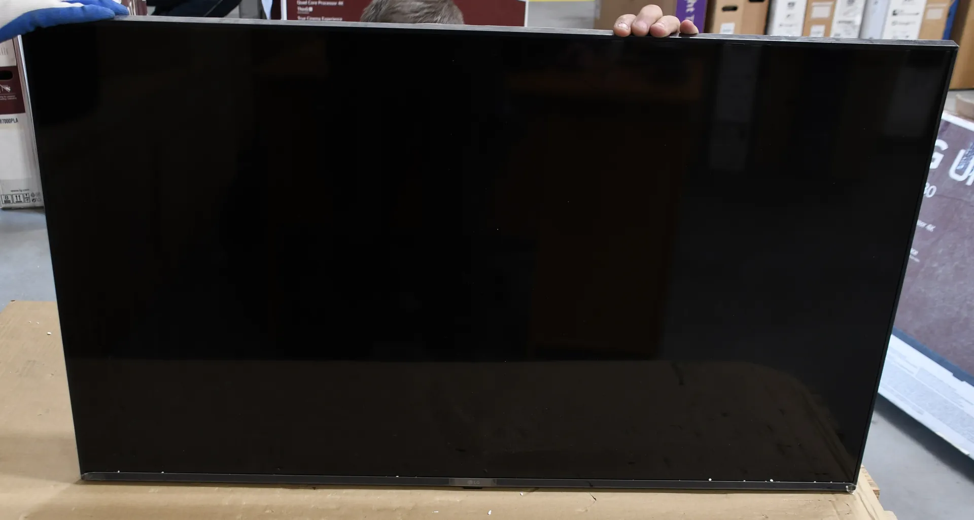 Zdjęcie przedstawiające czarny, wyłączony ekran telewizora o dużym formacie, który opiera się o kartonowe opakowanie. Na górnej krawędzi ekranu widoczne są dłonie osoby go trzymającej.