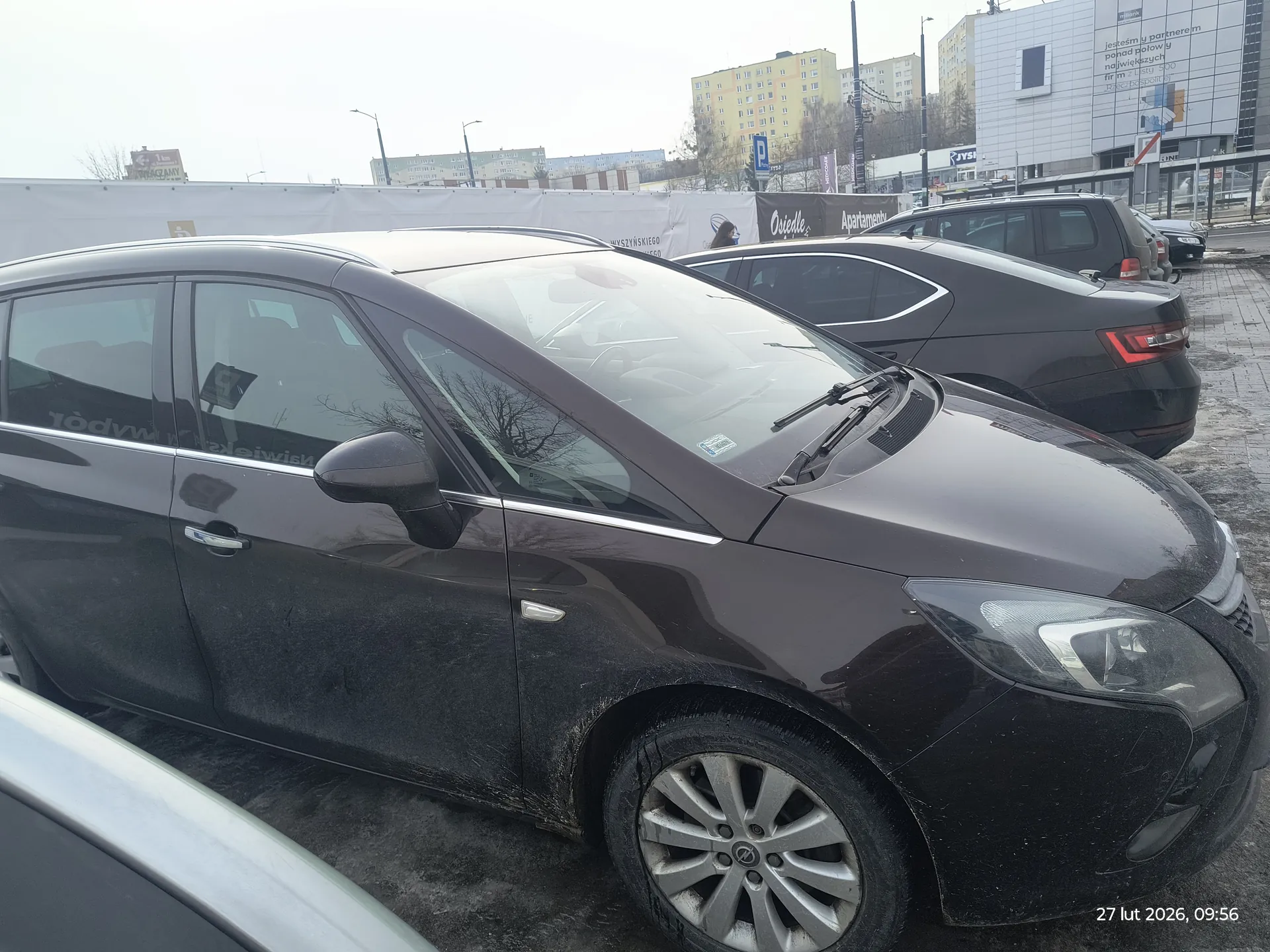 Stojące samochody na parkingu, na pierwszym planie ciemny samochód osobowy z widocznym kołem i lusterkiem bocznym. W tle inne pojazdy, białe barierki, budynki mieszkalne i biurowe z napisami 'Ośrodek', 'Apartamenty' oraz sklepem 'JYSK'. Na prawo od budynków znak parkingu (P). W prawym dolnym rogu widoczny znaczek daty i godziny: '27 lut 2026, 09:56'. Niebo zachmurzone, nawierzchnia parkingu mokra.