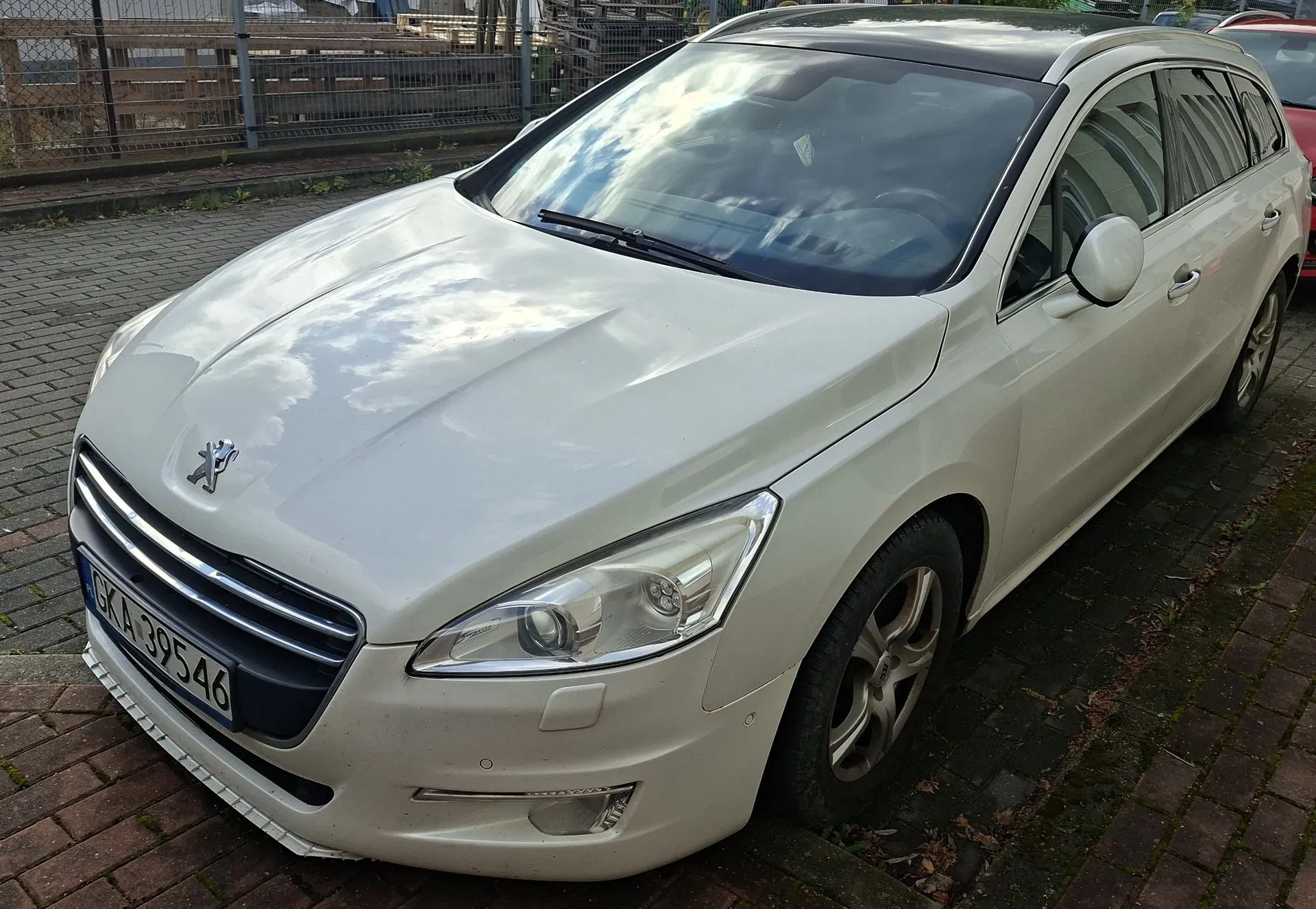 Biały samochód osobowy marki Peugeot w nadwoziu kombi, zaparkowany na kostce brukowej. Widoczny jest przód i prawy bok pojazdu. Samochód posiada tablicę rejestracyjną o numerze GKA 39546.
