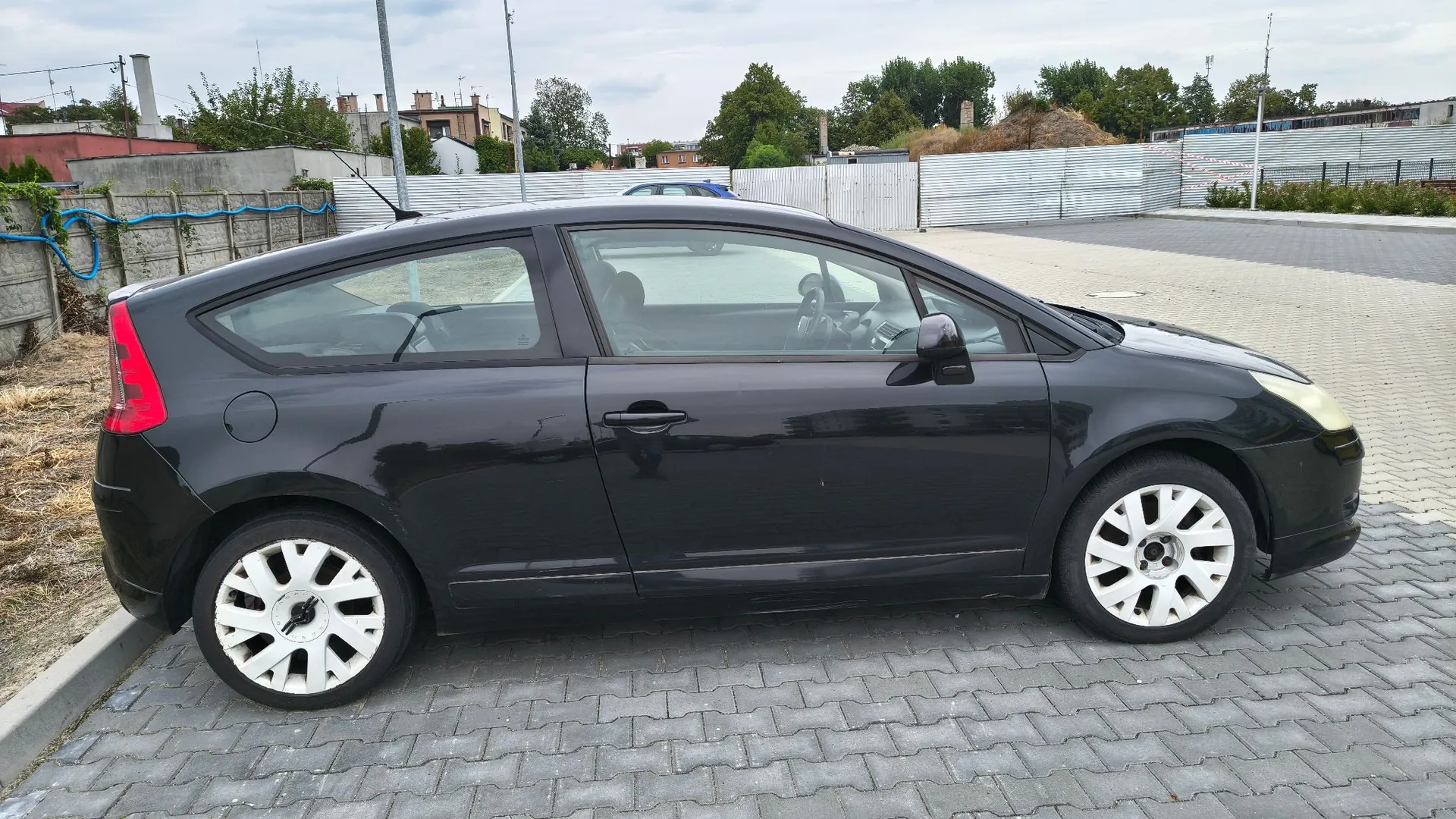 Czarny samochód osobowy typu hatchback zaparkowany bokiem na szarym bruku. Pojazd znajduje się na otwartym placu, w tle widoczne jest metalowe ogrodzenie, zielone drzewa oraz fragmenty budynków pod zachmurzonym niebem.