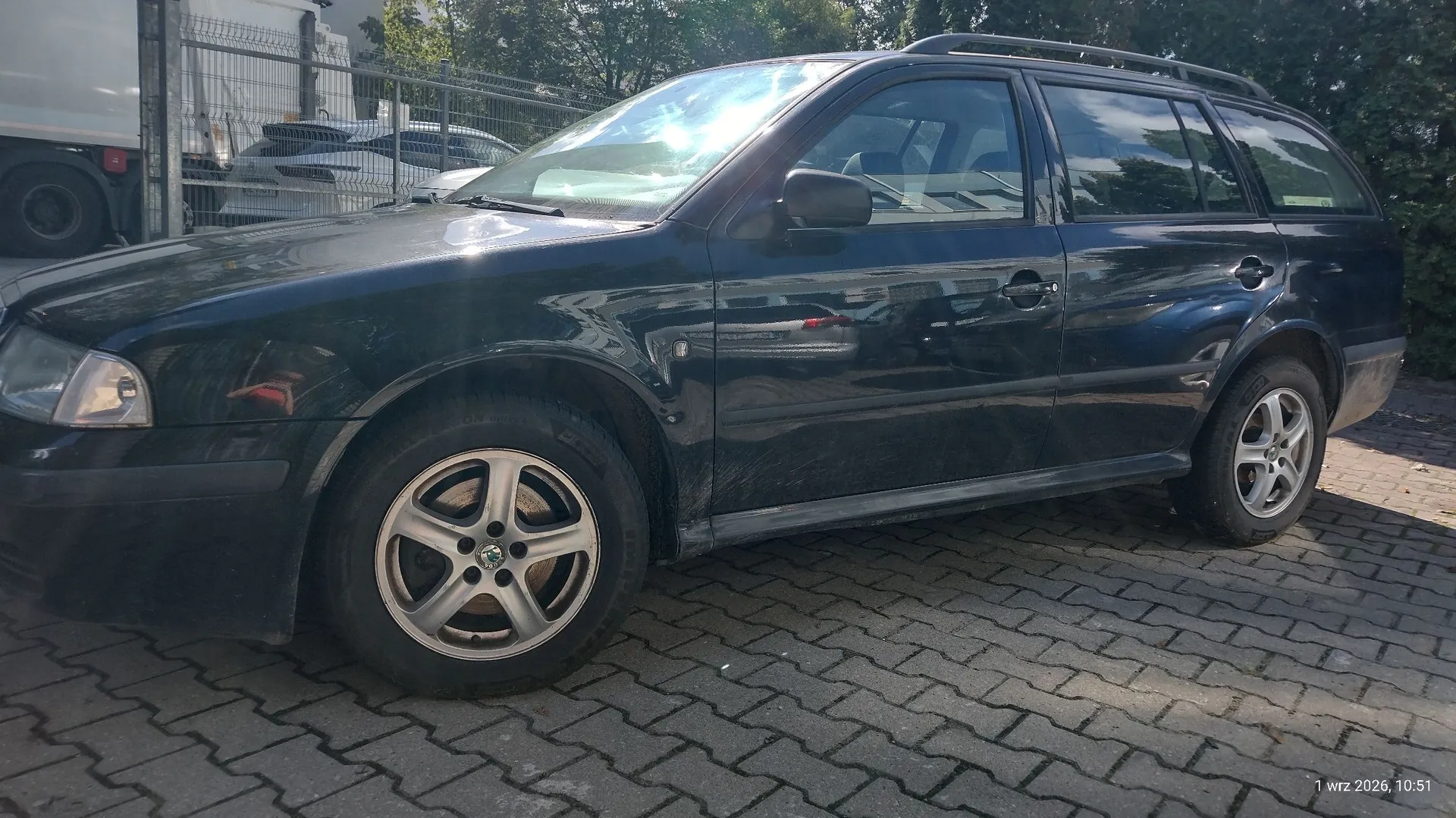 Czarny samochód osobowy typu kombi zaparkowany na kostce brukowej. Pojazd jest widoczny z profilu, z przodu i z boku. W tle widać ogrodzenie z siatki, białą ciężarówkę oraz zielone drzewa. W prawym dolnym rogu znajduje się data i godzina: 1 wrz 2021, 10:51.
