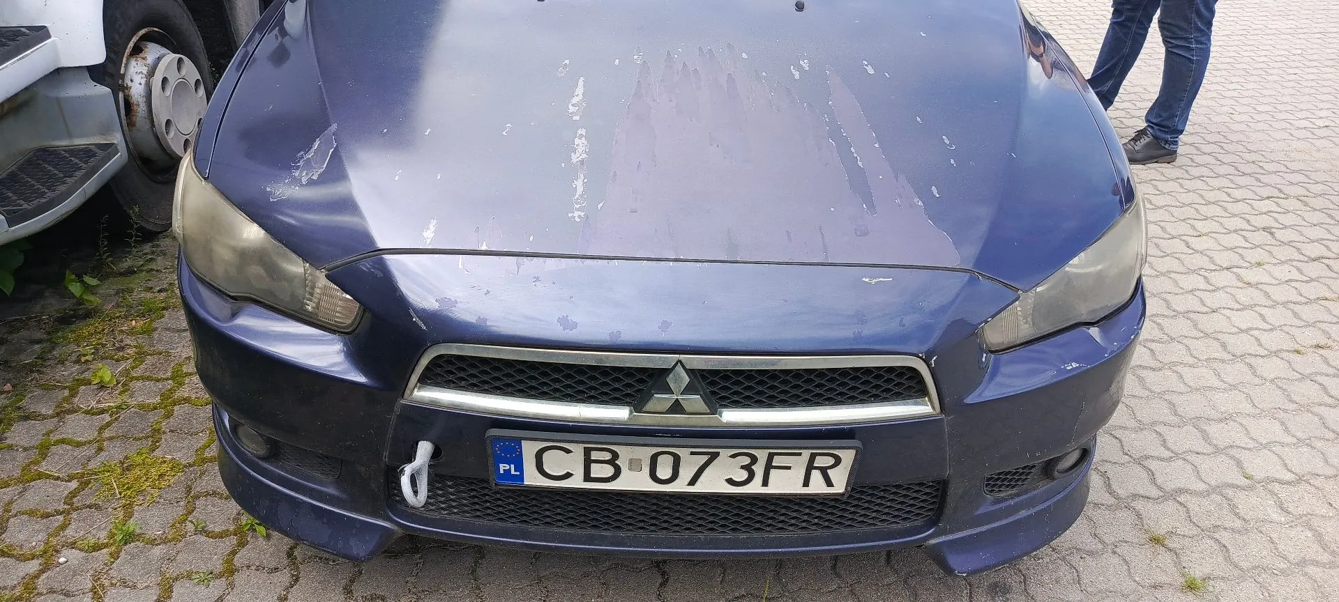 Przód granatowego samochodu marki Mitsubishi z widocznymi uszkodzeniami karoserii na masce i zderzaku. Samochód posiada polską tablicę rejestracyjną o numerze CB 073FR. Po lewej stronie widoczny jest fragment białego pojazdu.