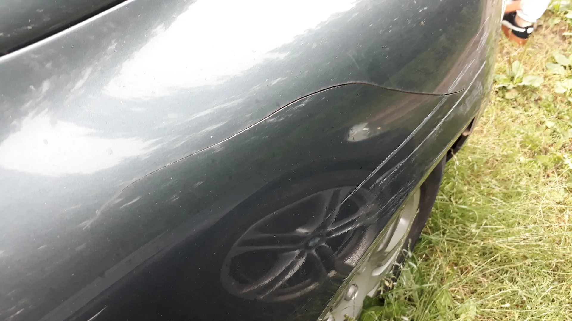 Zbliżenie na ciemnoszary lakier samochodu, pokazujące uszkodzenie w postaci pęknięcia lub szczeliny w elemencie nadwozia nad tylnym kołem. Na powierzchni lakieru widoczne są liczne drobne zarysowania. W tle widać fragment srebrnej felgi oraz zieloną trawę.