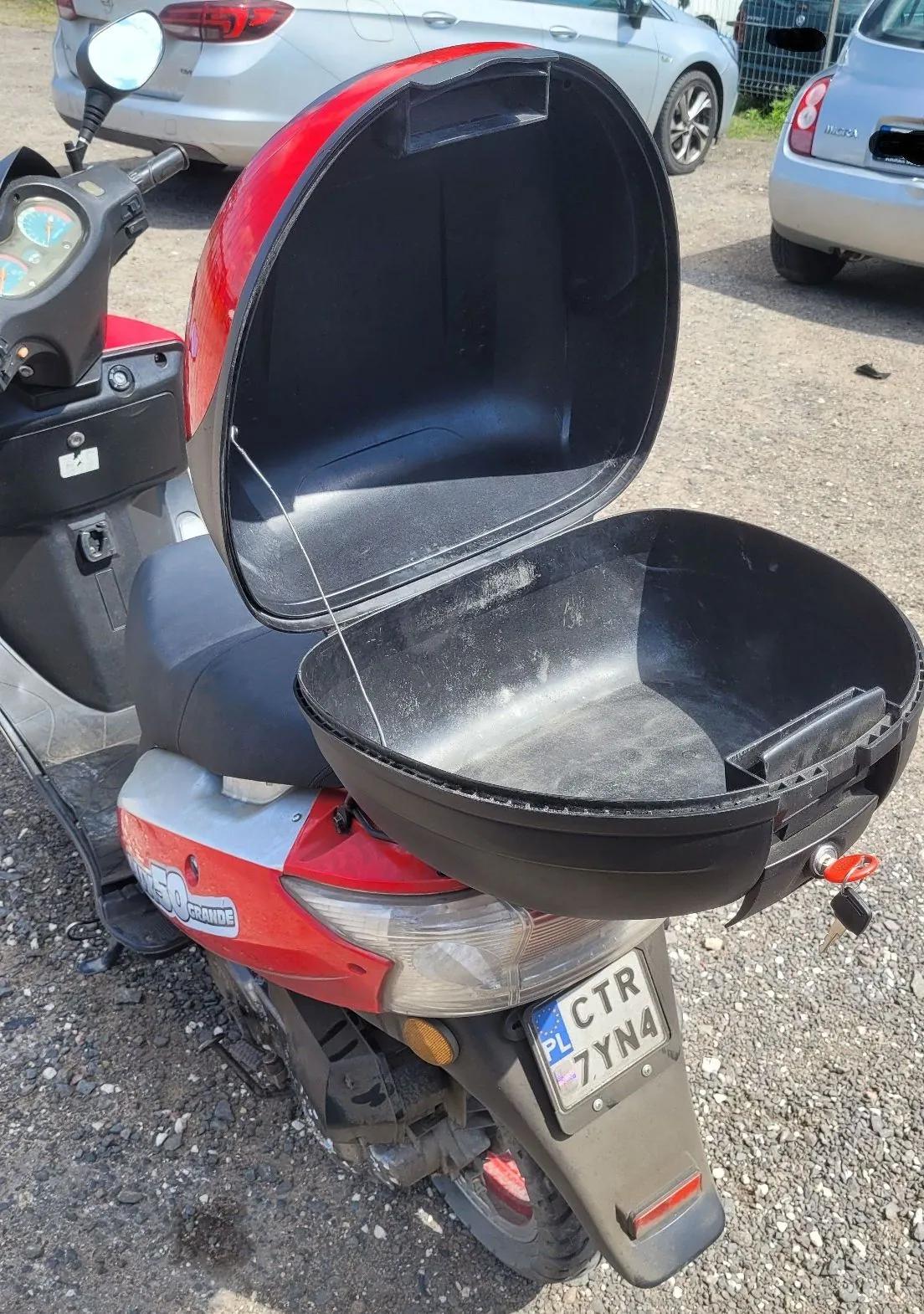 Czerwono-srebrny skuter zaparkowany na żwirowym podłożu. Z tyłu pojazdu znajduje się otwarta, czarna skrzynia bagażowa z czerwoną pokrywą. Na tylnym błotniku widoczna jest polska tablica rejestracyjna o numerze CTR 7YN4. W tle widoczne są fragmenty dwóch samochodów: srebrnego sedana oraz jasnego samochodu miejskiego.