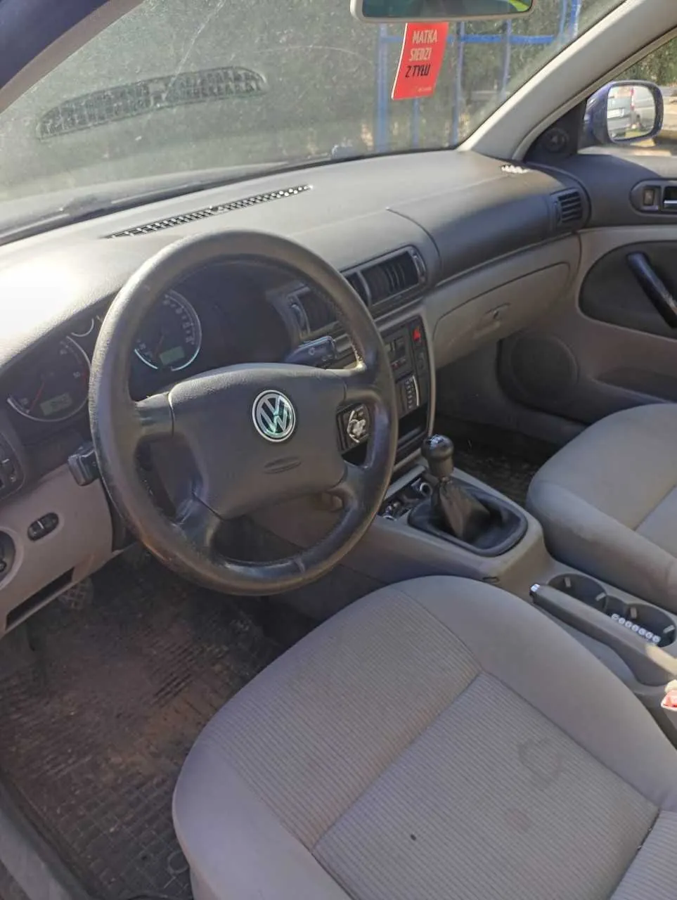 Wnętrze samochodu marki Volkswagen z kierownicą z logo VW, deską rozdzielczą z licznikami, dźwignią zmiany biegów i szarymi siedzeniami. Na przedniej szybie widoczny czerwony znak z napisem 'MATKA SIEDZI Z TYŁU'.