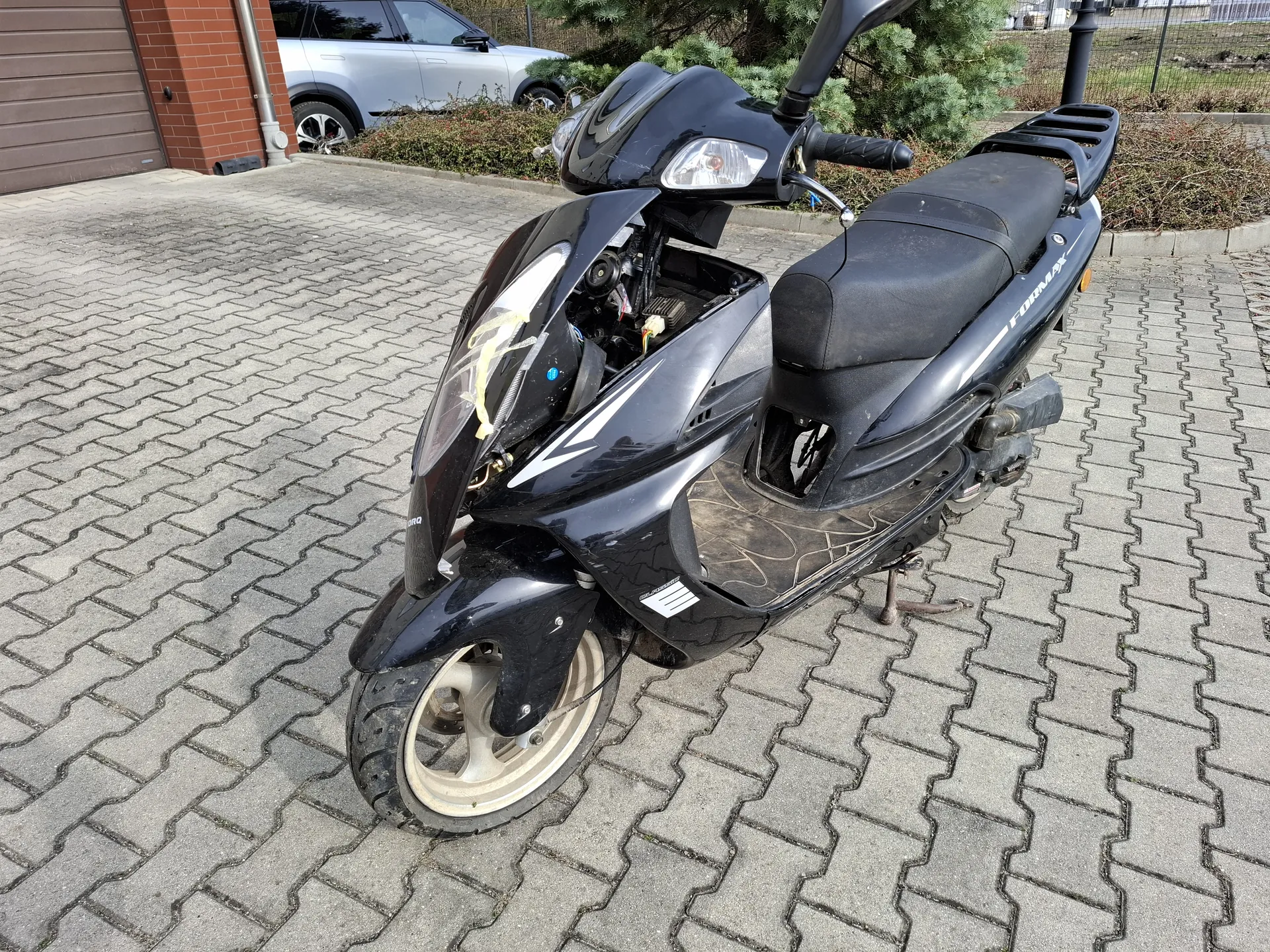 Czarny skuter marki Honda model Forza zaparkowany na brukowanej powierzchni. Przednia osłona skutera jest otwarta, odsłaniając elementy mechanizmu. Na boku skutera widoczny napis 'FORZA', a na przedniej części napis 'HONDA'. W tle srebrny samochód osobowy, ceglany budynek z brązowymi drzwiami garażowymi, krzewy i ogrodzenie. Tekst na skuterze jest czytelny i poziomo ustawiony.