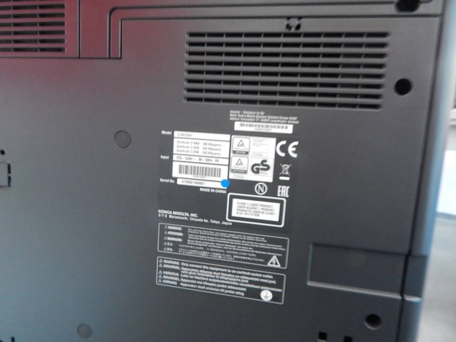 Zdjęcie tylnej obudowy urządzenia elektronicznej, prawdopodobnie drukarki lub kserokopiarki. Widoczna jest czarna, plastikowa obudowa z kratkami wentylacyjnymi oraz naklejka z informacjami technicznymi. Na naklejce znajduje się tekst: 'Model C302301', 'Input 220-240V ~ 50-60Hz 6A', 'Serial No. A7R0021009921', 'MADE IN CHINA' oraz nazwa producenta 'KONICA MINOLTA, INC. 2-7-2, Higashikawasaki, Chiyoda-ku, Tokyo, Japan'. Widoczne są również znaki certyfikacji, takie jak CE, GS oraz EAC.