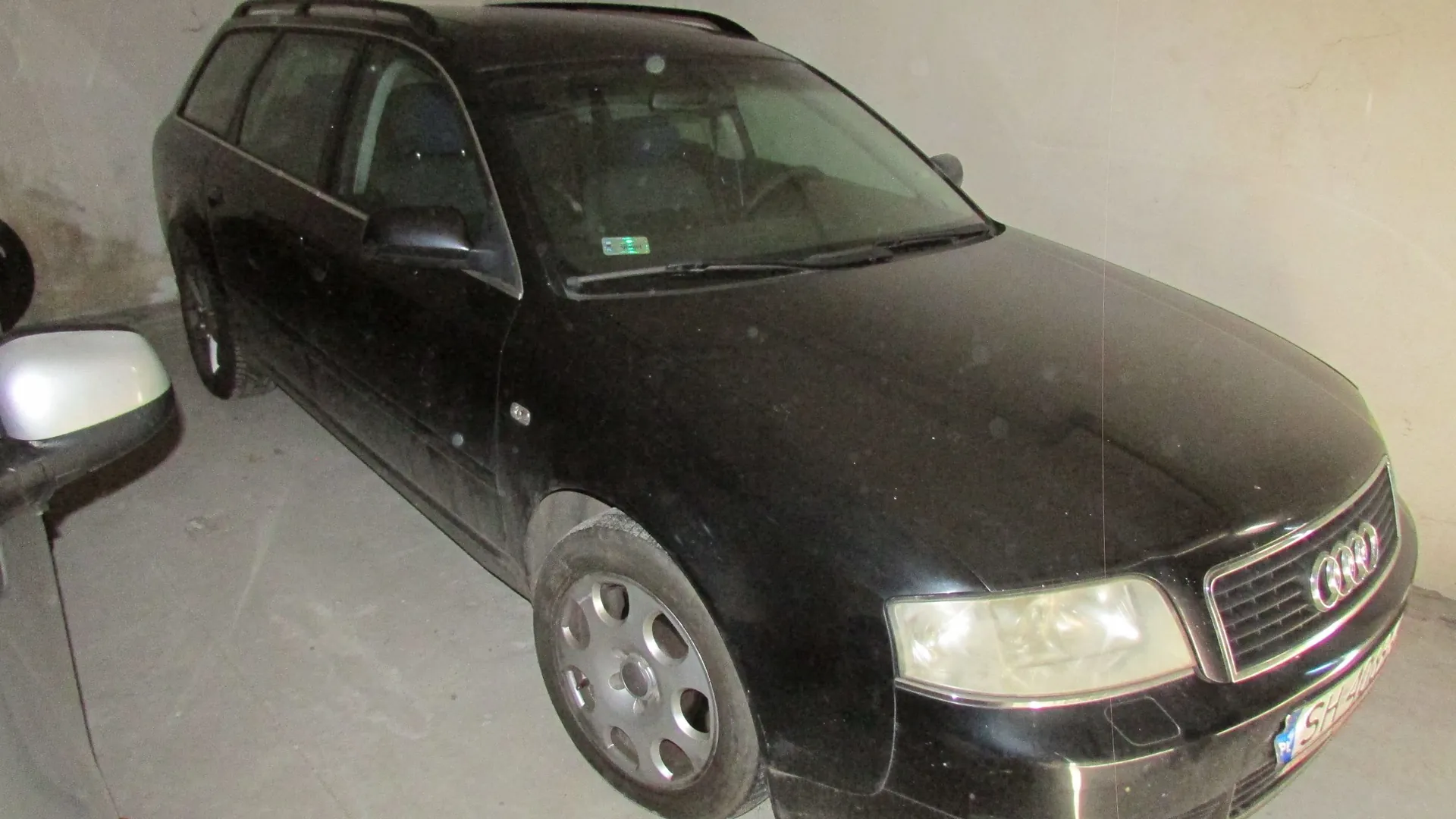 Czarny samochód marki Audi w nadwoziu kombi, zaparkowany w jasnym garażu lub pomieszczeniu. Pojazd jest zakurzony, widoczne są reflektory, grill z logo Audi oraz fragment tablicy rejestracyjnej z literami SH. Po lewej stronie kadru widoczny jest fragment lusterka innego pojazdu.