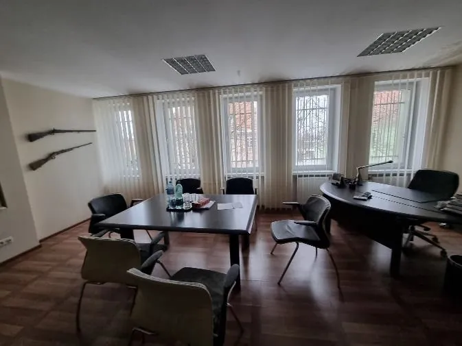 Biuro z ciemnym stołem konferencyjnym w centrum, na którym znajdują się butelka wody, dokumenty i naczynia. Wokół stołu stoją czarne i szare krzesła. Po prawej stronie znajduje się zakrzywione biurko z lampą, książkami i telefonem. Na lewej ścianie wiszą dwa karabiny. Pokój ma podłogę z drewnianych płytek, jasne ściany oraz cztery okna z pionowymi roletami, przez które widać zewnętrzne drzewa i budynki. Na suficie znajdują się kwadratowe oprawy oświetleniowe.