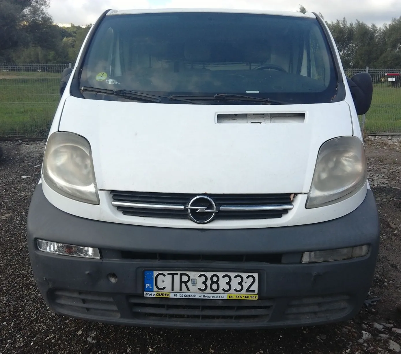 Widok z przodu białego samochodu marki Opel. Pojazd ma szary plastikowy zderzak z zamontowaną polską tablicą rejestracyjną o numerze CTR 38332. Poniżej numeru rejestracyjnego znajduje się żółty pasek z tekstem: 'Moto GUREK 87-122 Grodzisk ul. Kowalewska 45 tel. 515 168 902'. Reflektory są lekko zmatowiałe. W tle widoczny jest zielony płot i drzewa pod zachmurzonym niebem.