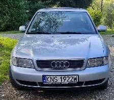 Srebrny samochód marki Audi widziany od przodu, zaparkowany na żwirowym podłożu na tle zieleni. Pojazd posiada tablicę rejestracyjną o numerze KNS 1922W.