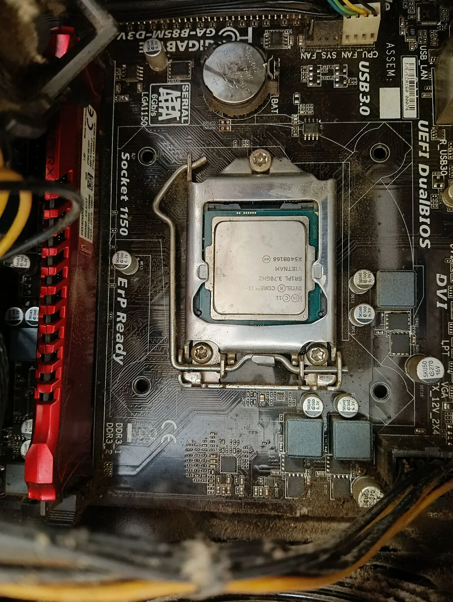 Zbliżenie na płytę główną komputera z zainstalowanym procesorem w gnieździe Socket 1150. Na procesorze widnieje napis: 'INTEL CORE i3-4130 VIETNAM SRIPL 3.70GHz X5408168'. Płyta główna jest czarna, z widocznymi napisami 'Gigabyte', 'USB 3.0', 'ErP Ready' oraz 'UEFI DualBIOS'. Po lewej stronie widoczny jest czerwony element obudowy lub chłodzenia, a na dole i po prawej stronie przebiegają żółto-czarne kable zasilające.