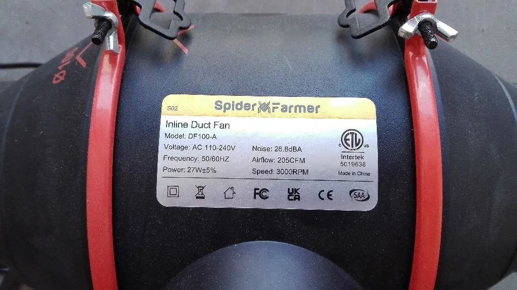 Zbliżenie na czarny wentylator kanałowy z czerwonymi klamrami mocującymi. Na obudowie znajduje się biała etykieta z napisem 'Spider Farmer' oraz specyfikacją techniczną: 'S02 Inline Duct Fan, Model: DF100-A, Voltage: AC 110-240V, Frequency: 50/60HZ, Power: 27W±5%, Noise: 26.8dBA, Airflow: 205CFM, Speed: 3000RPM'. Na etykiecie widoczne są również certyfikaty: ETL Intertek, FC, CE oraz SAA.